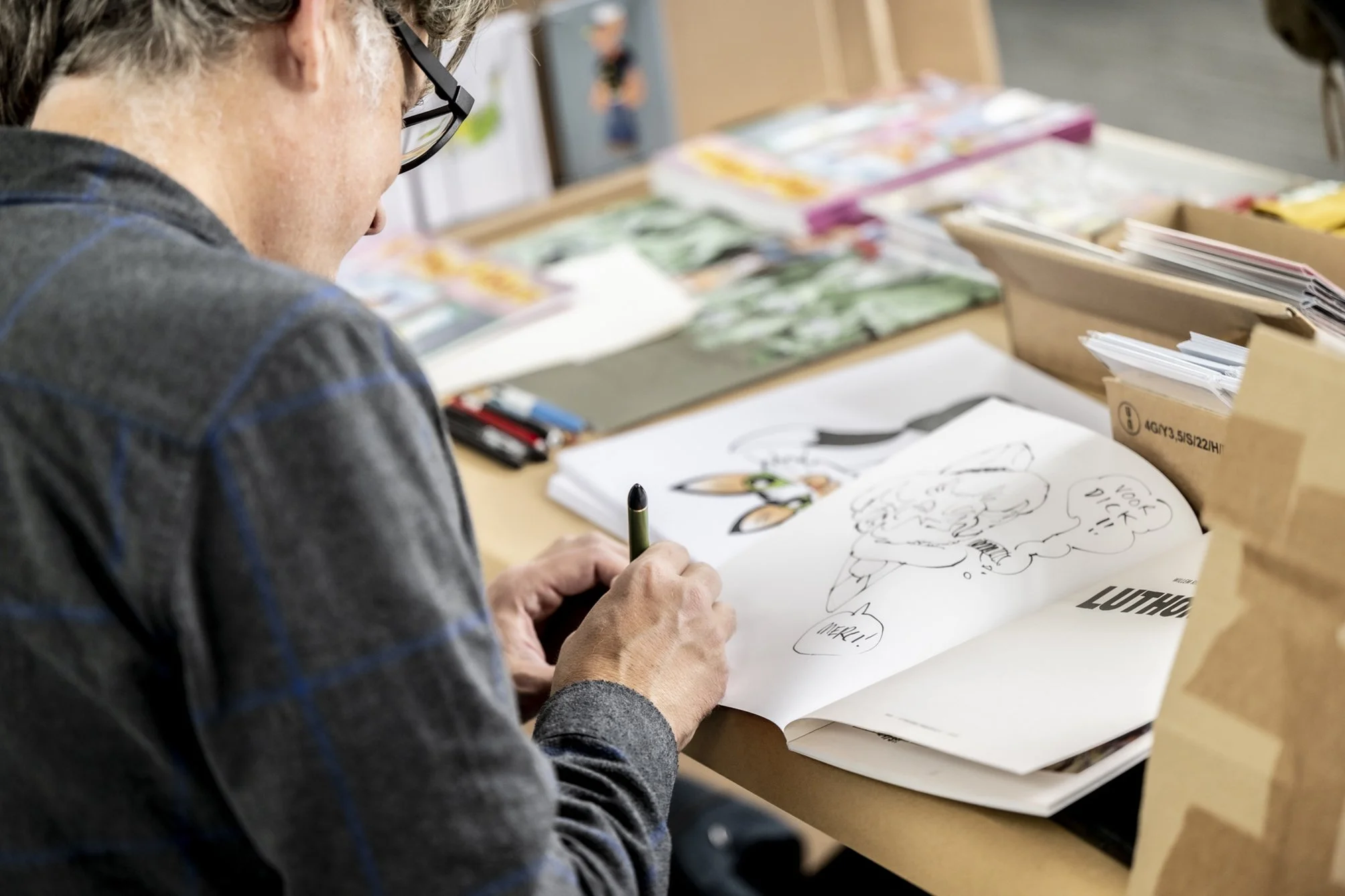 Persoon die een schets maakt op een grote blad met cartoon figuur en tekst, omringd door stapels papier en tekenmaterialen.