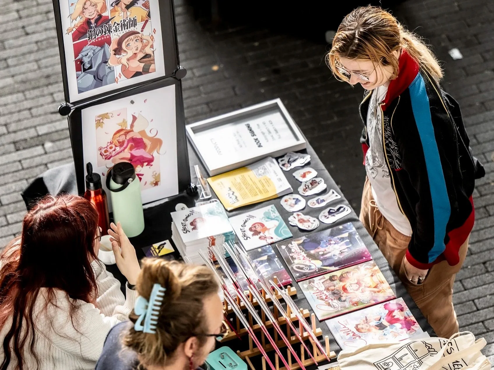 Twee mensen bekijken merchandise en kunst van anime of manga op een stand, met posters en stickers op de tafel.