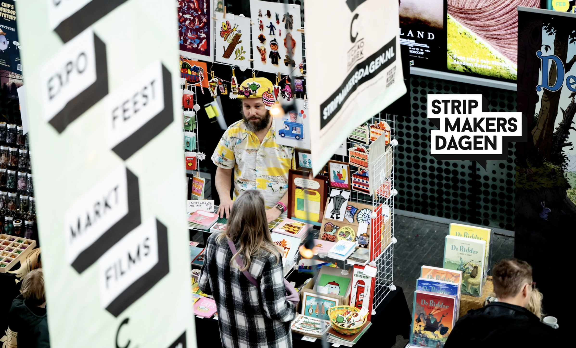Een binnenmarkt met een boekenkraam vol stripboeken en merchandise, mensen die naar de kraam kijken, decoraties aan de muur en grote borden met teksten zoals 'Strip Makers Dagen'.