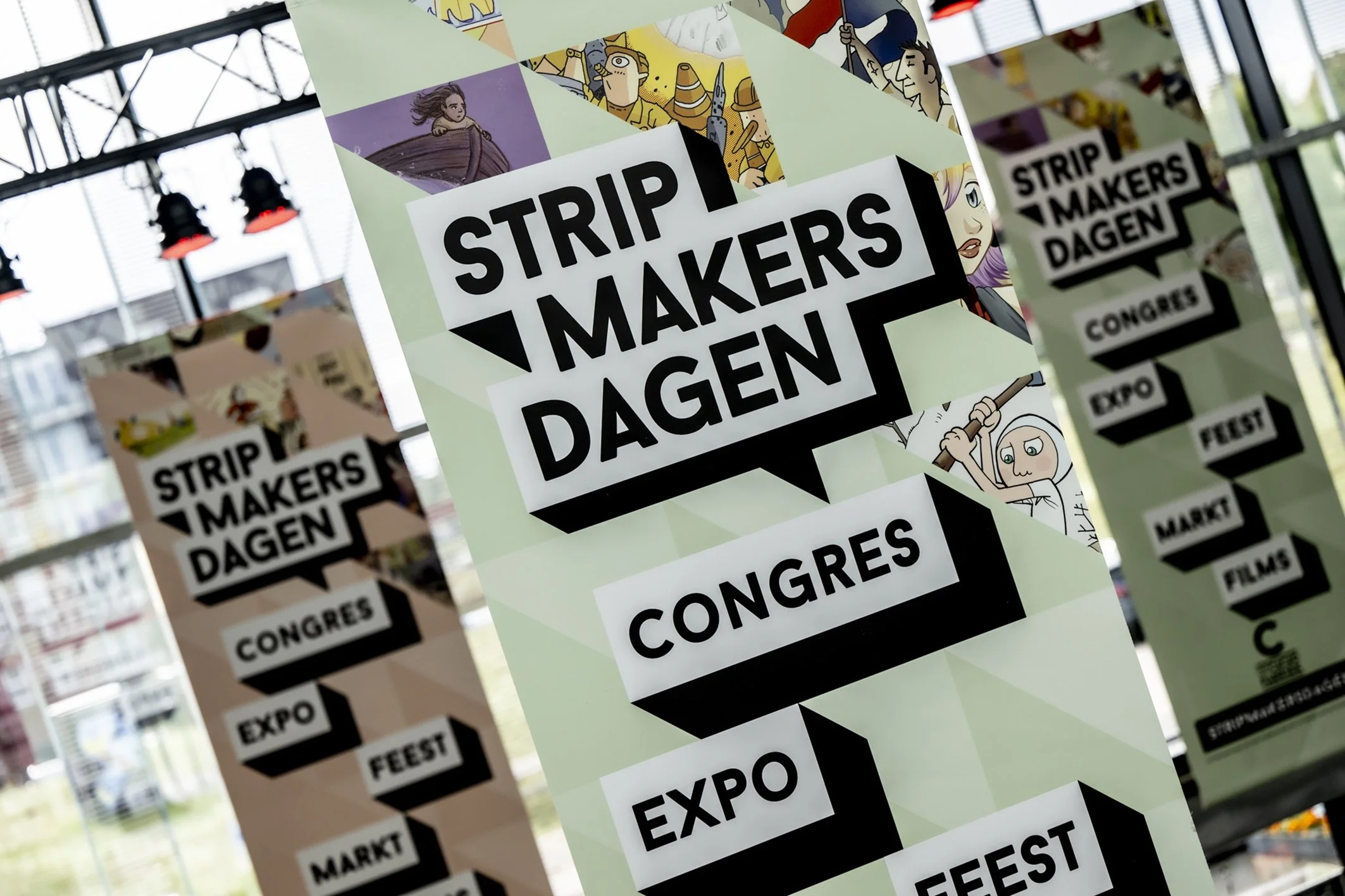 Vrijdragende posters met tekst 'Stripmakers DAGEN' en verschillende illustraties op een glazen muur.