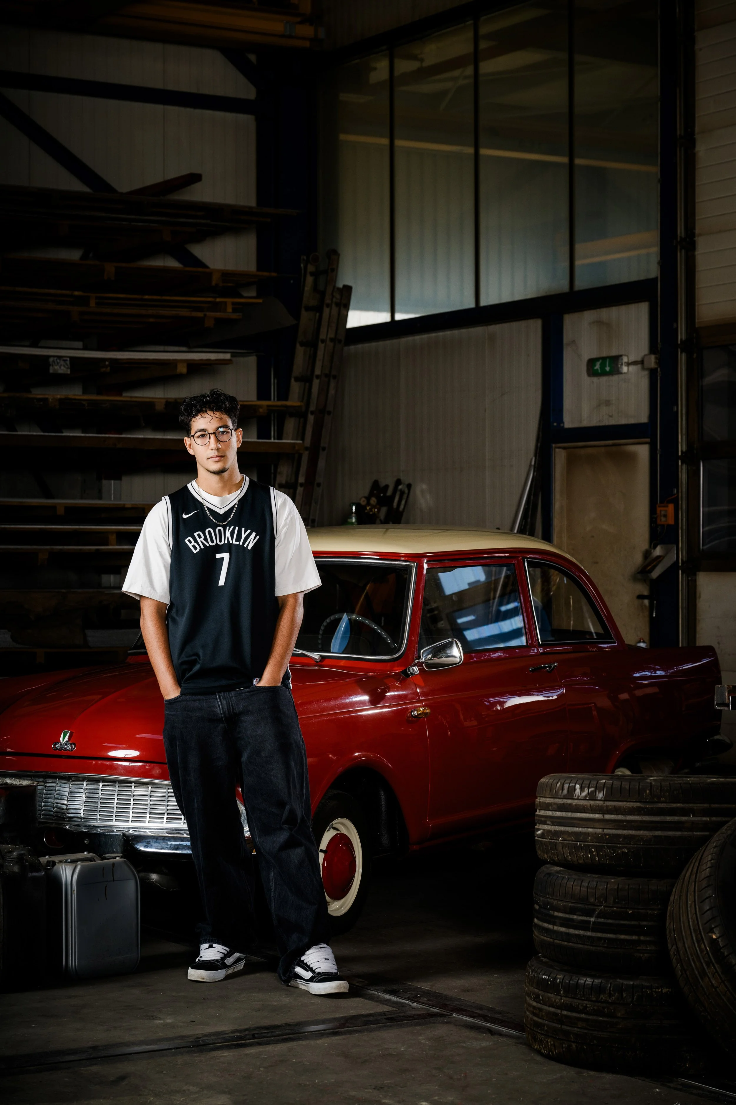 Junger Mann steht in einem Lagerraum mit alten Reifen, einem schwarzen Koffer, einem klassischen roten Auto und Holzregalen im Hintergrund. Er trägt eine Brooklyn-Jersey mit der Nummer 7, schwarze Jeans und weiße Turnschuhe.