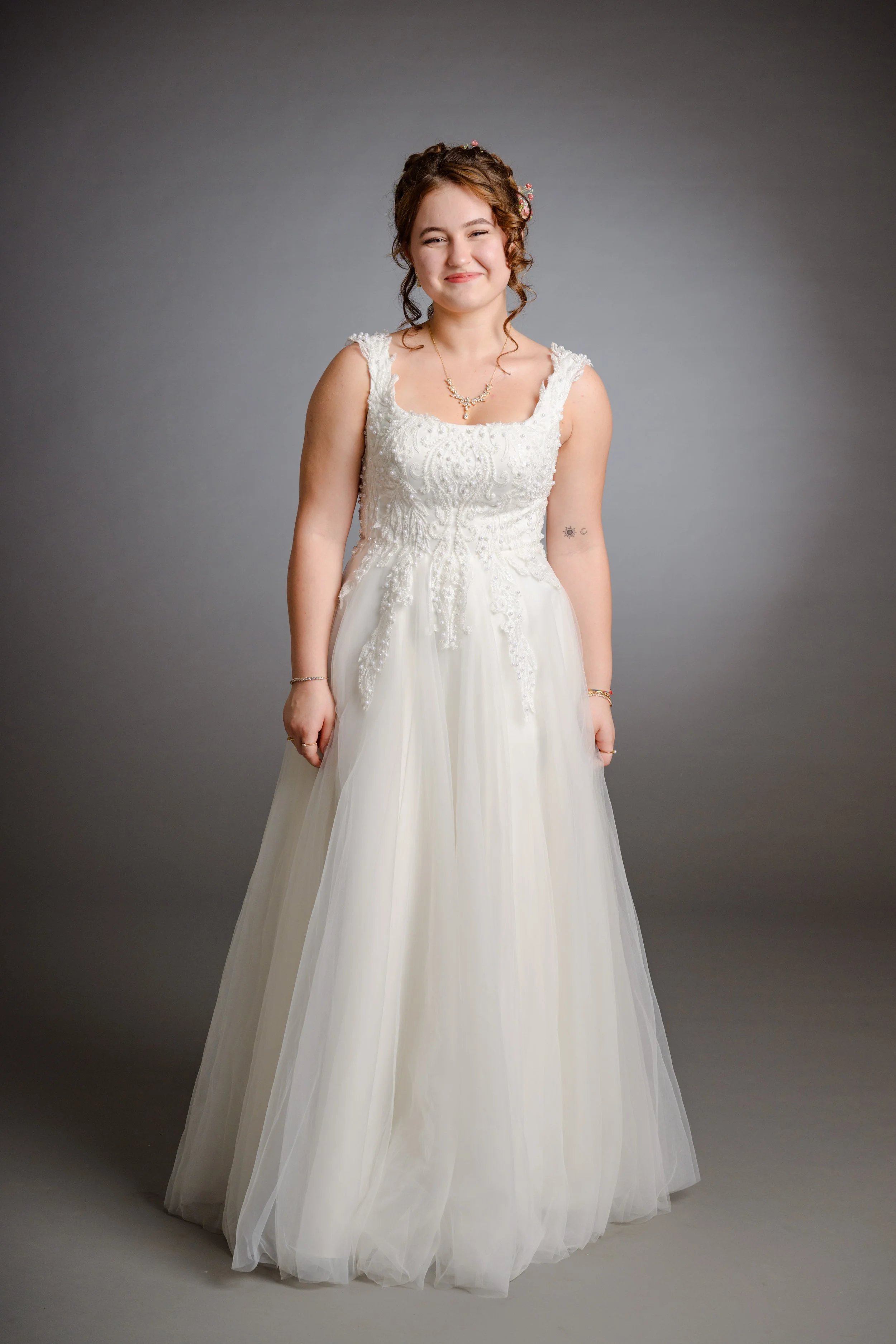 Eine junge Frau trägt ein weißes Hochzeitskleid mit Stickereien, steht vor grauem Hintergrund, lächelt leicht, trägt Schmuck und hat braune, lockige Haare.