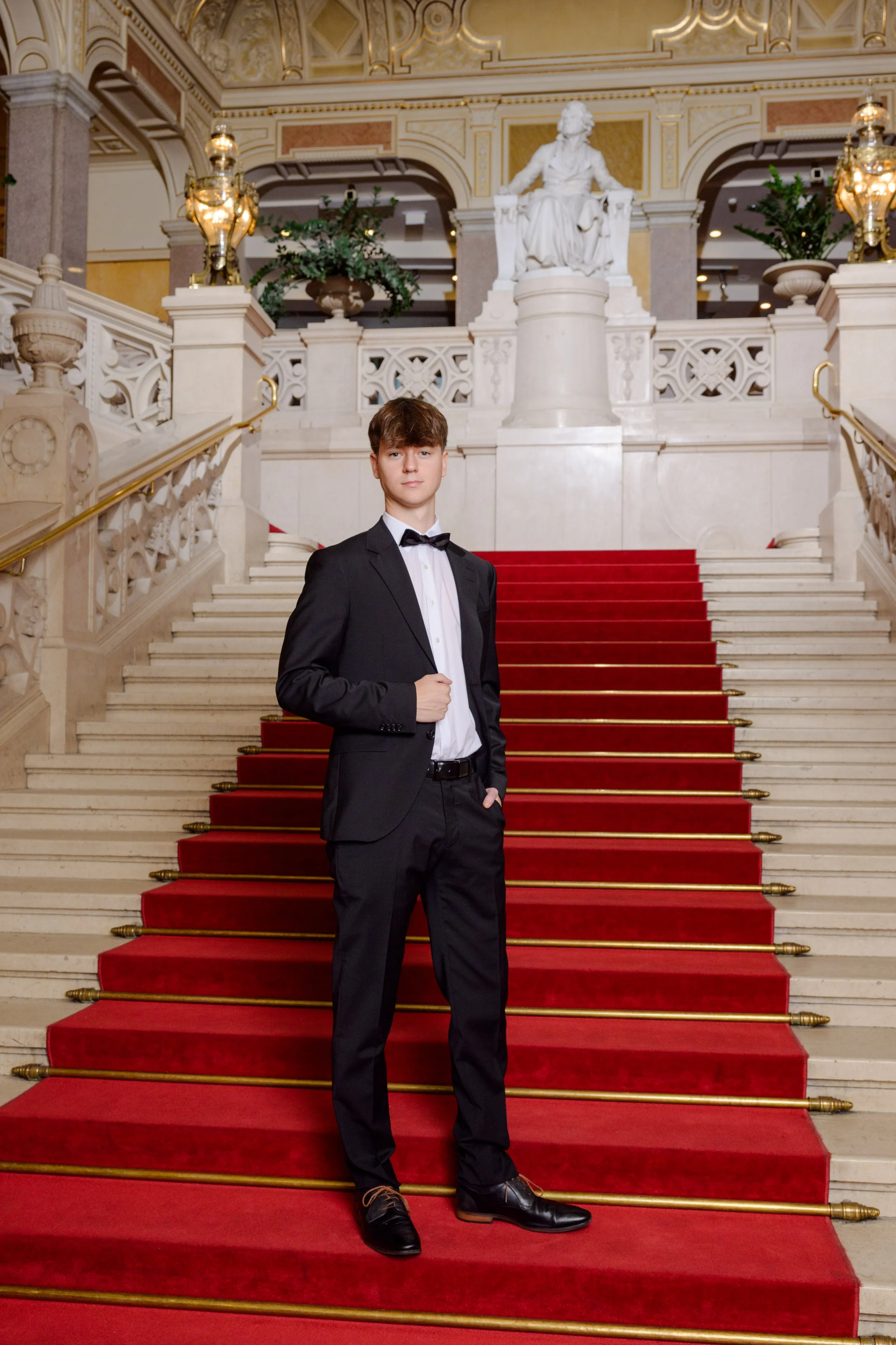 Junge in Smoking steht auf einer roten Treppe in einem prunkvollen, goldverzierten Saal mit Statue einer sitzenden Frau im Hintergrund.