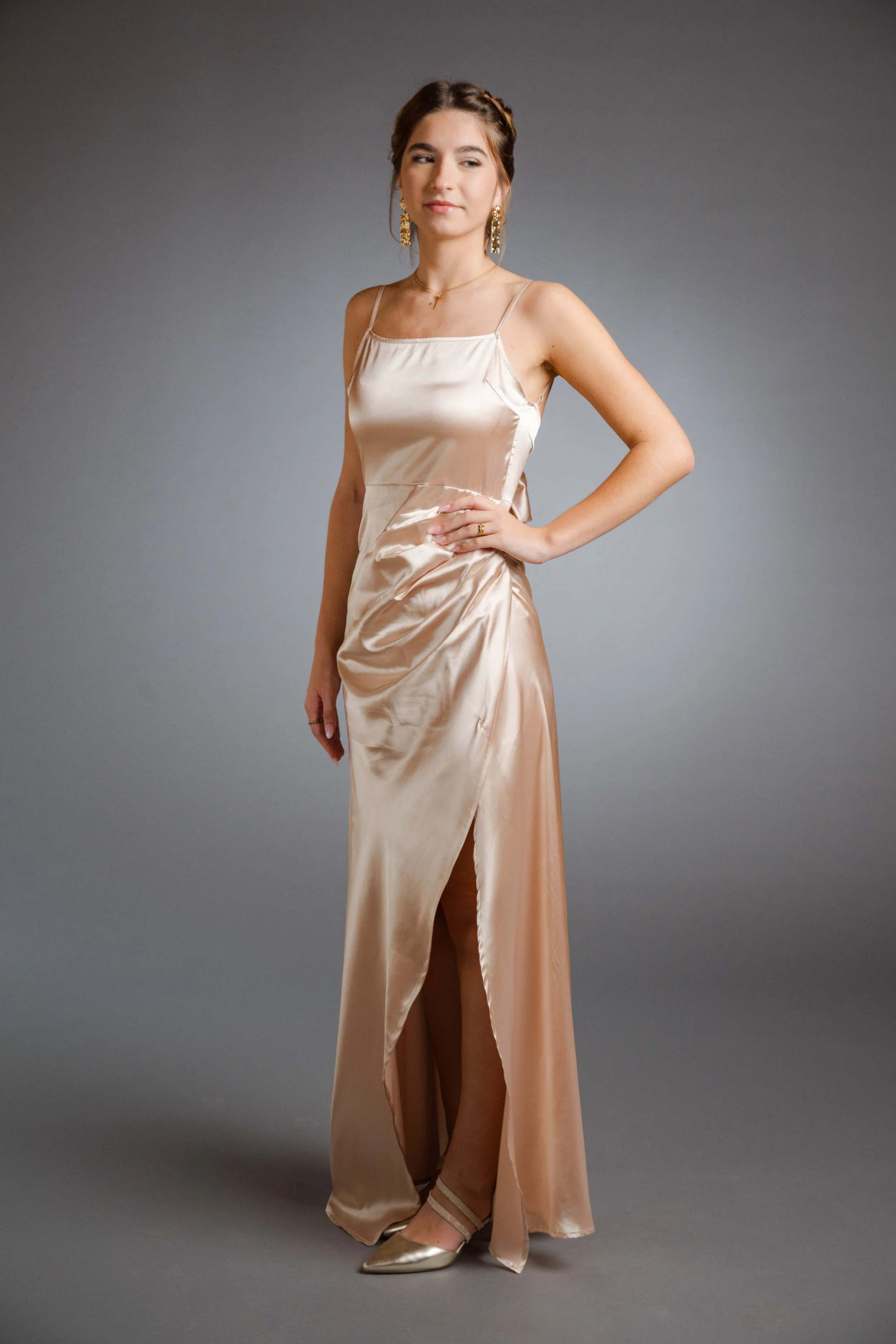 Junge Frau in einem seidigen, champagnefarbenen Abendkleid mit Spaghettiträgern und Schlitz, steht vor grauem Hintergrund.