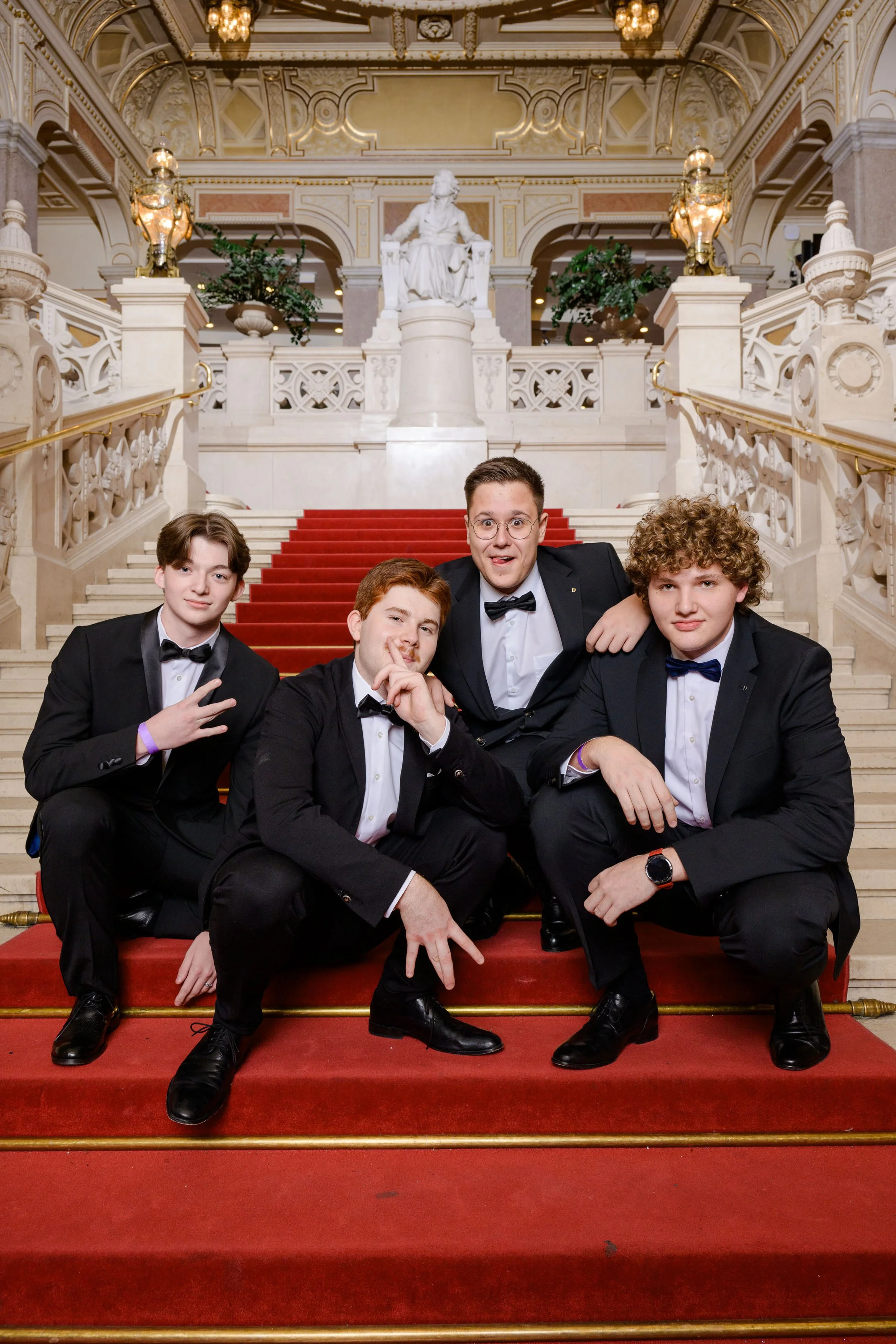 Vier junge Männer in formeller Kleidung sitzen auf einer Treppe in einem prunkvollen, historischen Gebäude mit Statuen und kunstvollen Dekorationen.