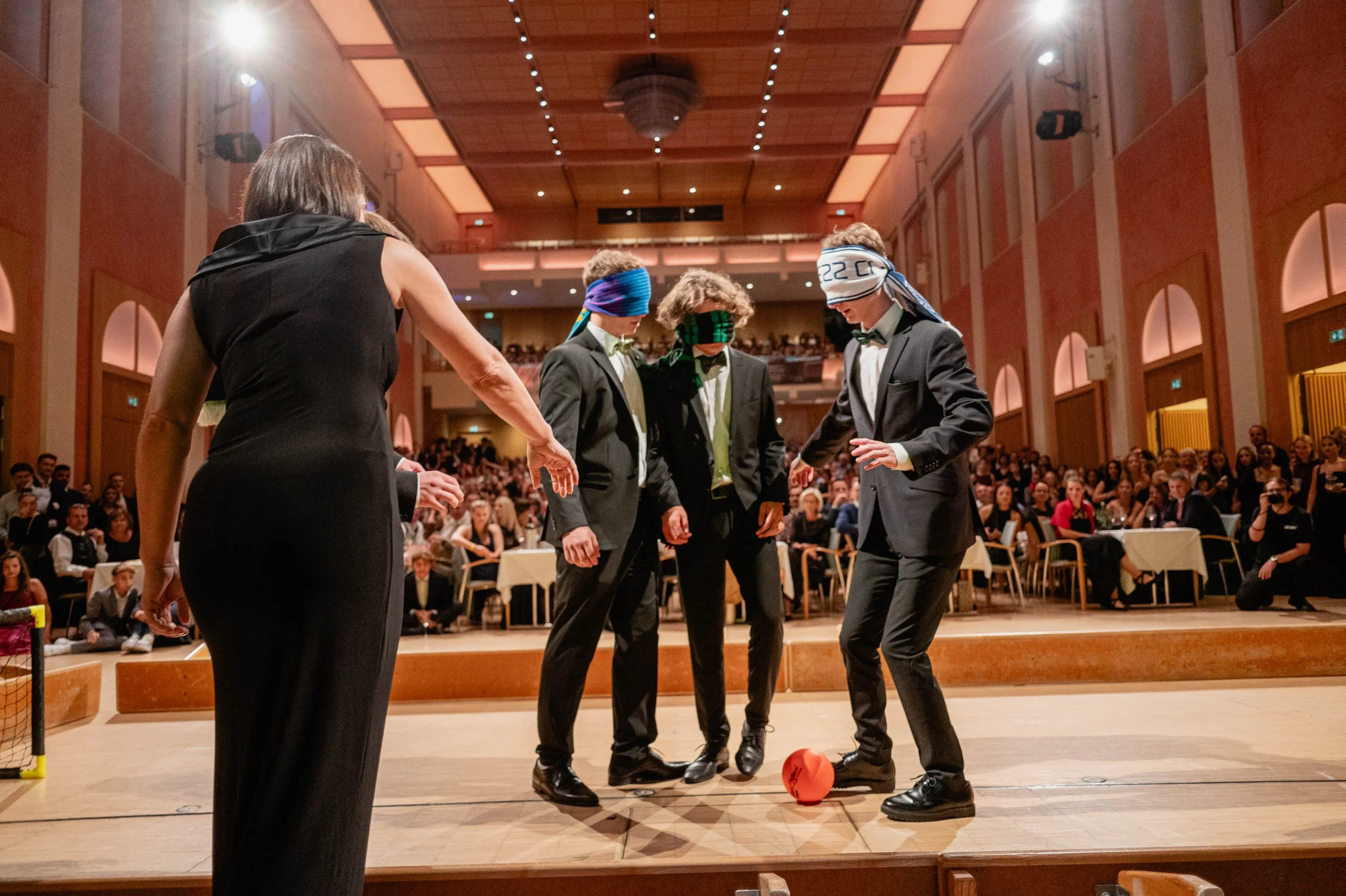 Vier Personen in formeller Kleidung, drei mit blauen und einem mit weißen Kopfbedeckungen, spielen mit einem roten Ball auf einer Bühne vor einem großen Publikum in einem festlichen Saal.