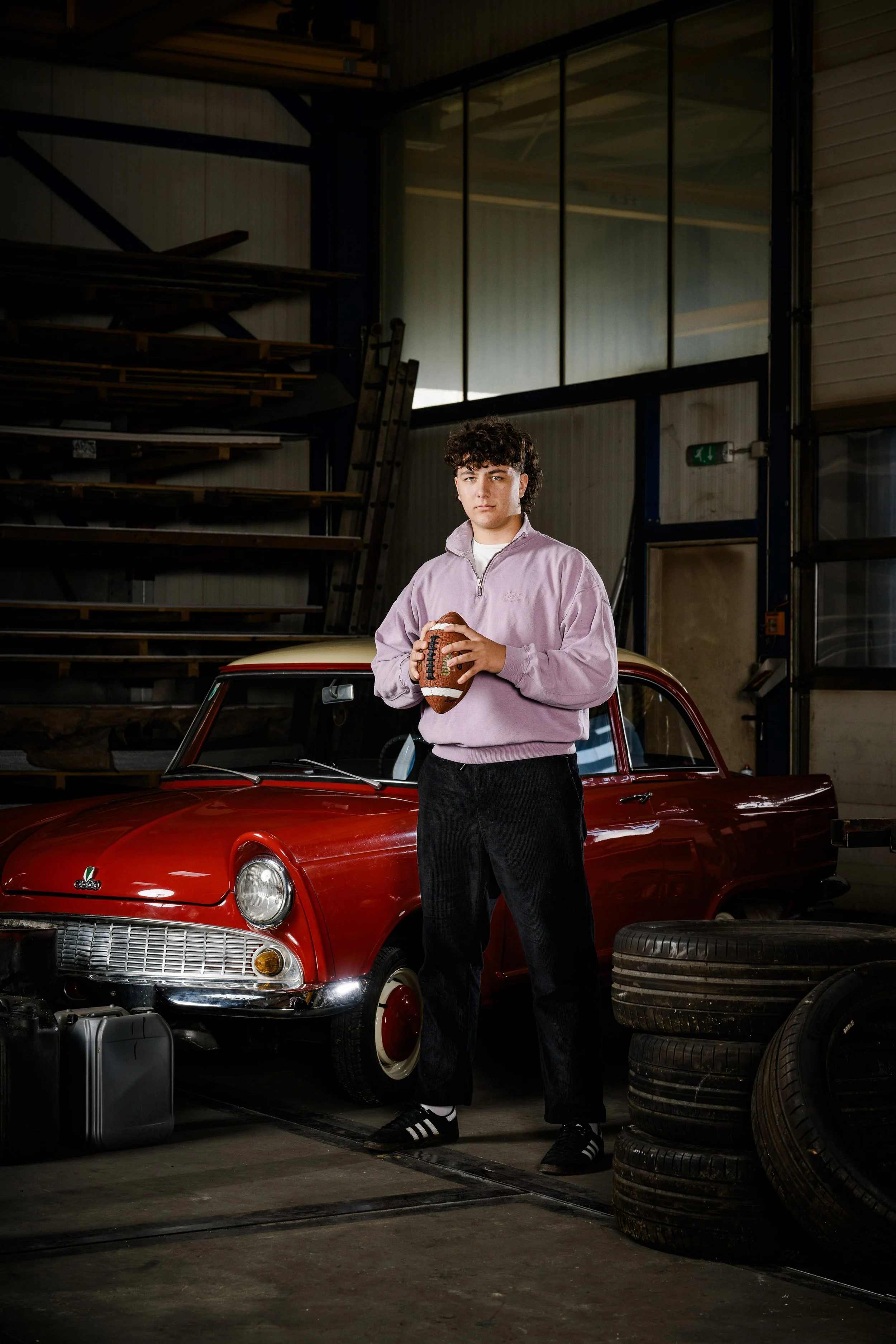 Junge hält american football vor einem roten Oldtimer in einer Werkstatt mit Reifen und Holzregalen.