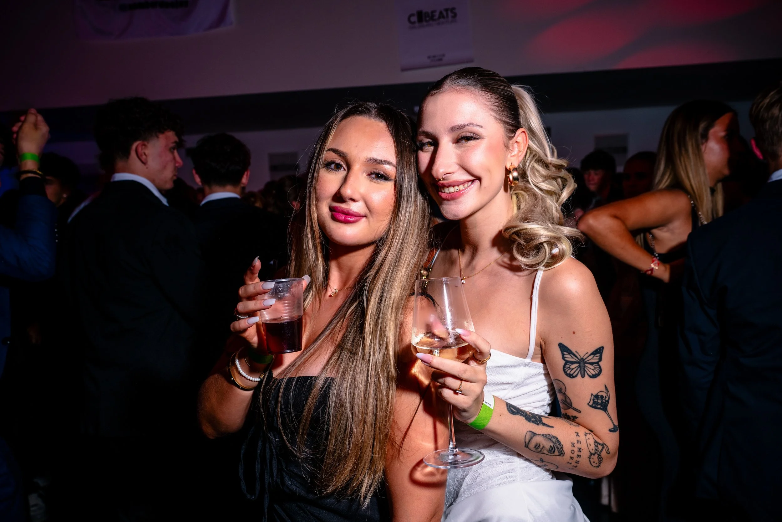Zwei junge Frauen bei einer Party, die in die Kamera lächeln, während sie Getränke halten, umgeben von weiteren Partygästen im Hintergrund.