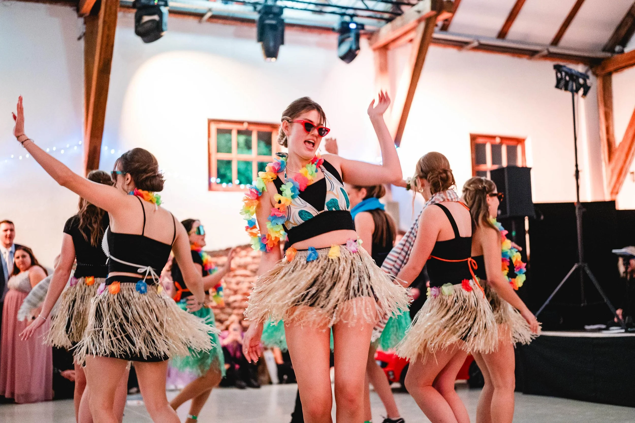 Mehrere junge Frauen tanzen auf einer Party, tragen Hula-Hoop-Kleider aus Stroh, bunte Blumen-Halsketten und Sonnenbrillen, im Hintergrund sind andere Gäste und eine Musikpult zu sehen.