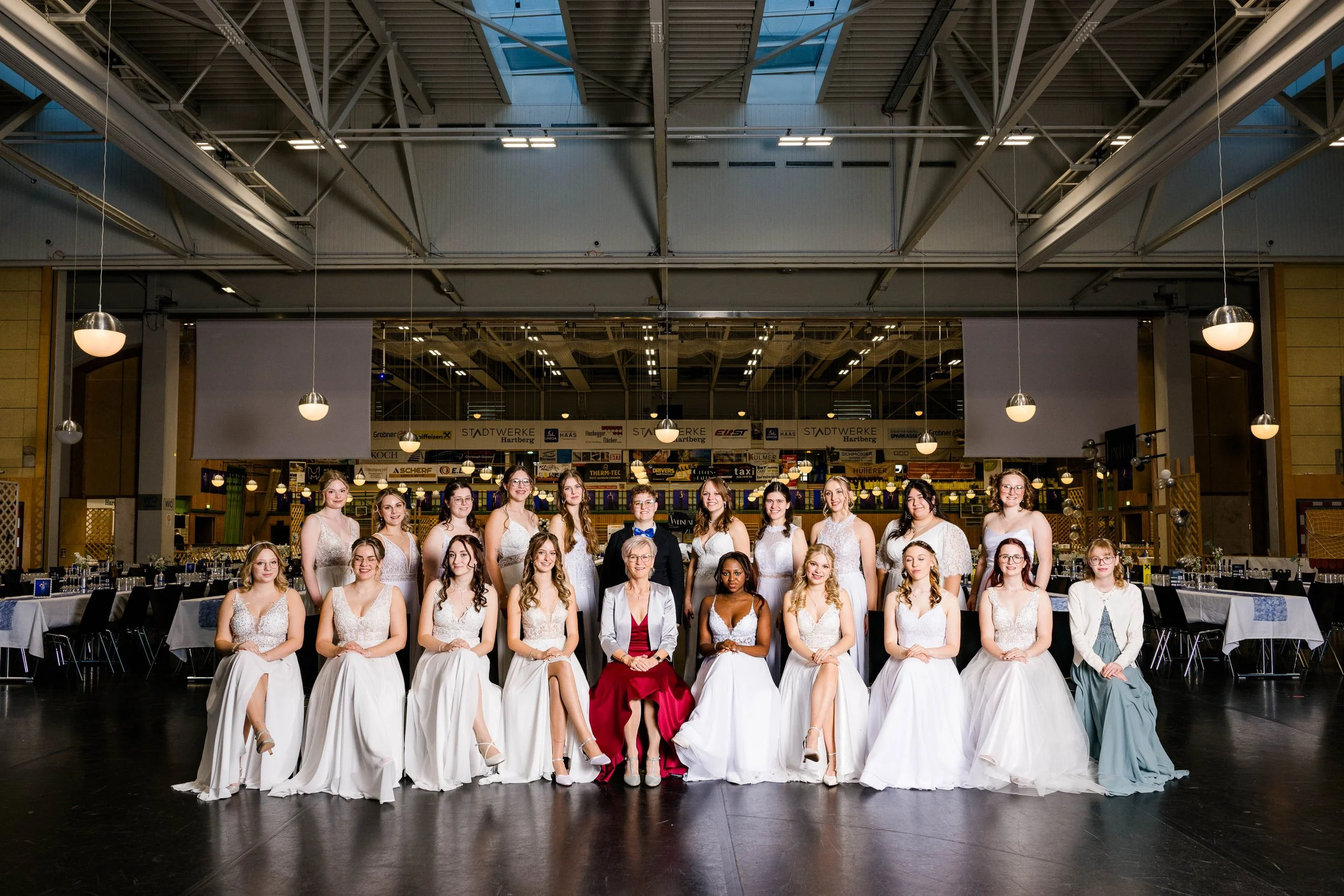 Eine Gruppe junger Frauen in formellen weißen Kleidern und zwei ältere Frauen in formeller Kleidung posieren für ein Gruppenfoto in einem großen Veranstaltungsraum.