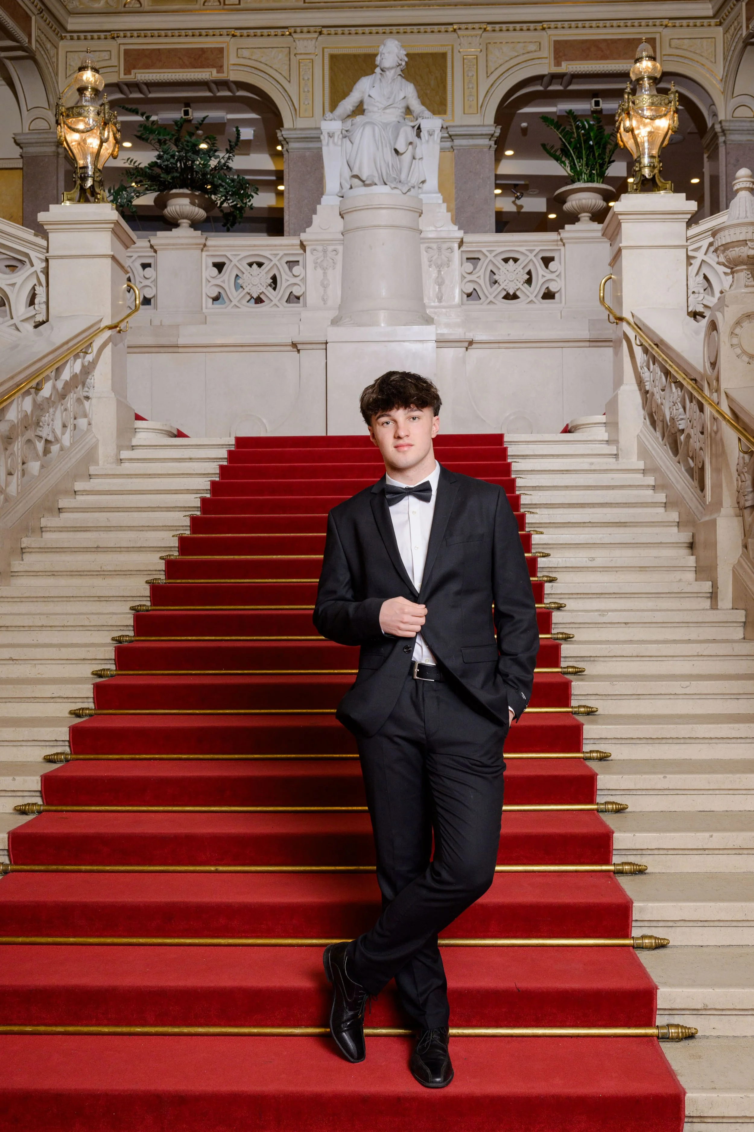 Ein junger Mann in einem schwarzen Anzug und Fliege steht an einer Treppe mit rotem Teppich, im Hintergrund eine weiße Statue und eine elegant dekorierte Halle.