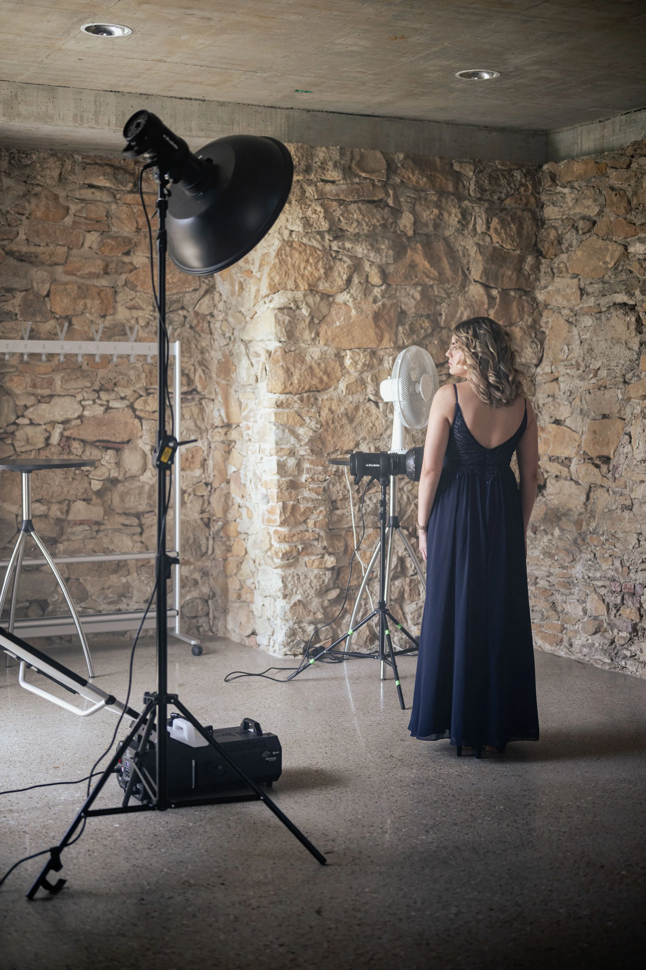 Eine Frau in einem dunklen Abendkleid steht in einem Raum mit Steinwänden und schaut zur Seite.