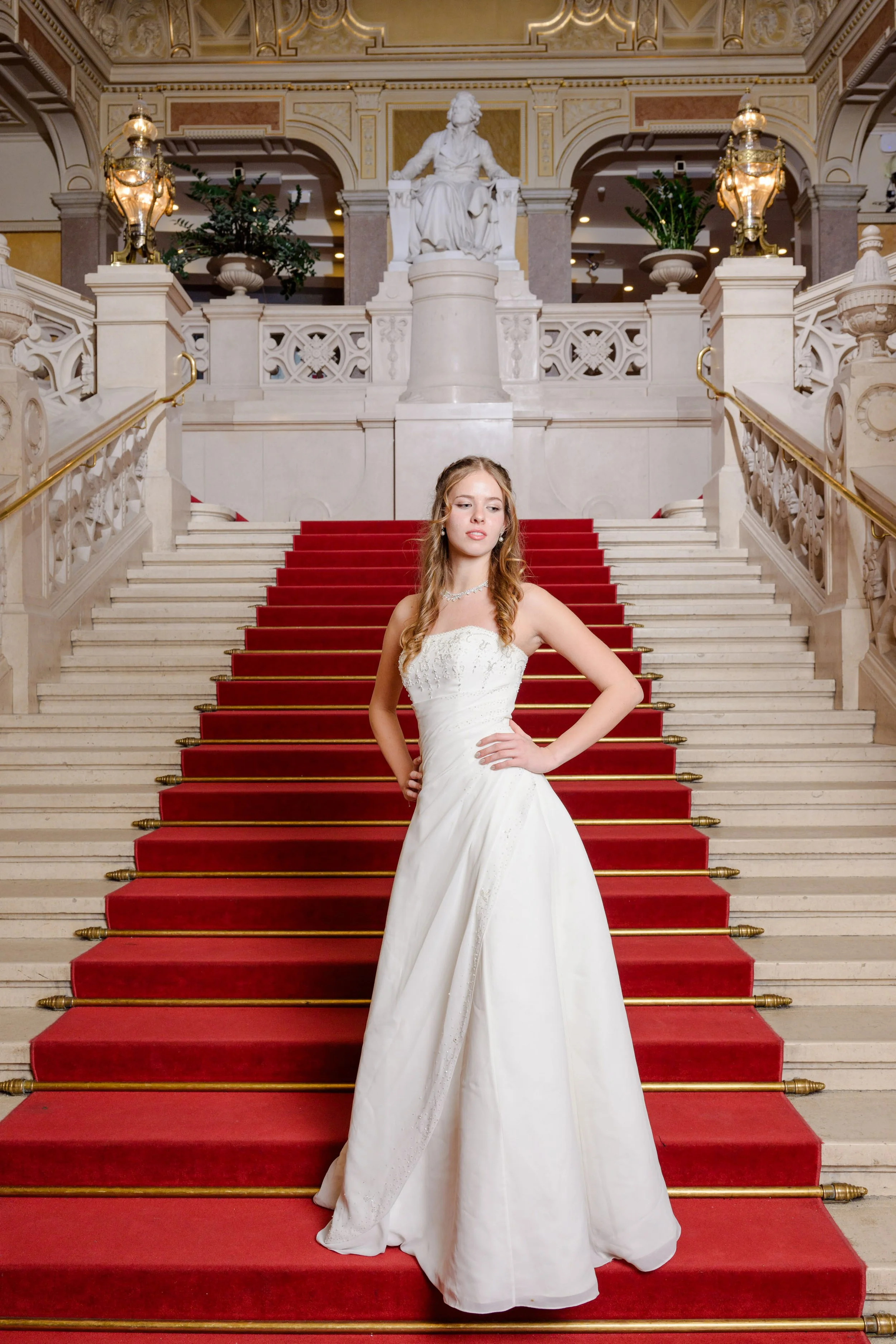 Junge Frau in einem weißen Brautkleid steht auf einer roten Treppe in einem prunkvollen Gebäude mit Goldverzierungen und Statue im Hintergrund.