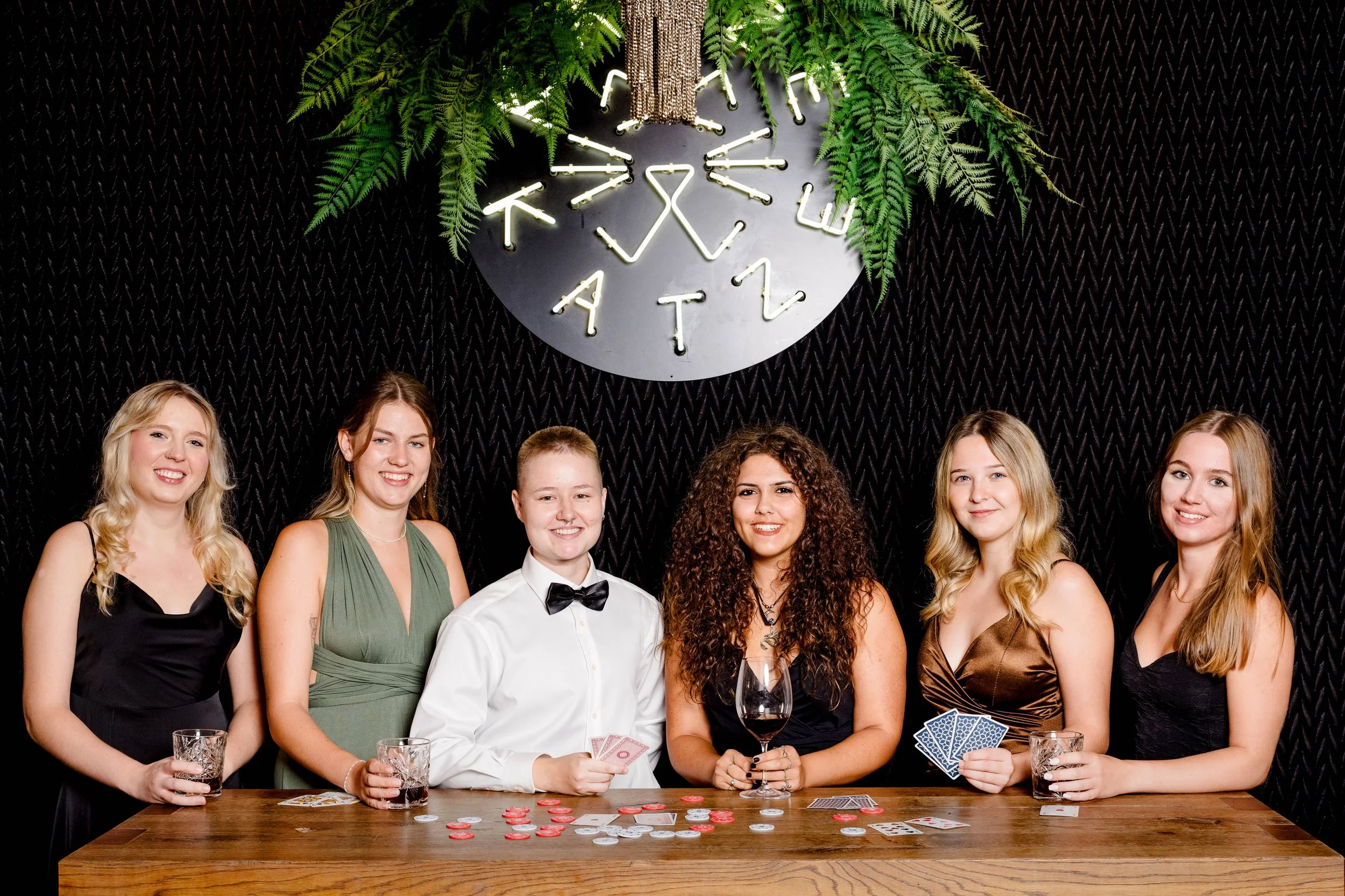 Sechs Frauen stehen hinter einem Holz-Tisch mit Spielkarten, Pokerchips und Gläsern, während sie eine Poker- oder Casino-Party feiern. Im Hintergrund eine schwarze Wand mit einer leuchtenden Markierung, die Wörter wie 'CATT' und eine Vielzahl von grü