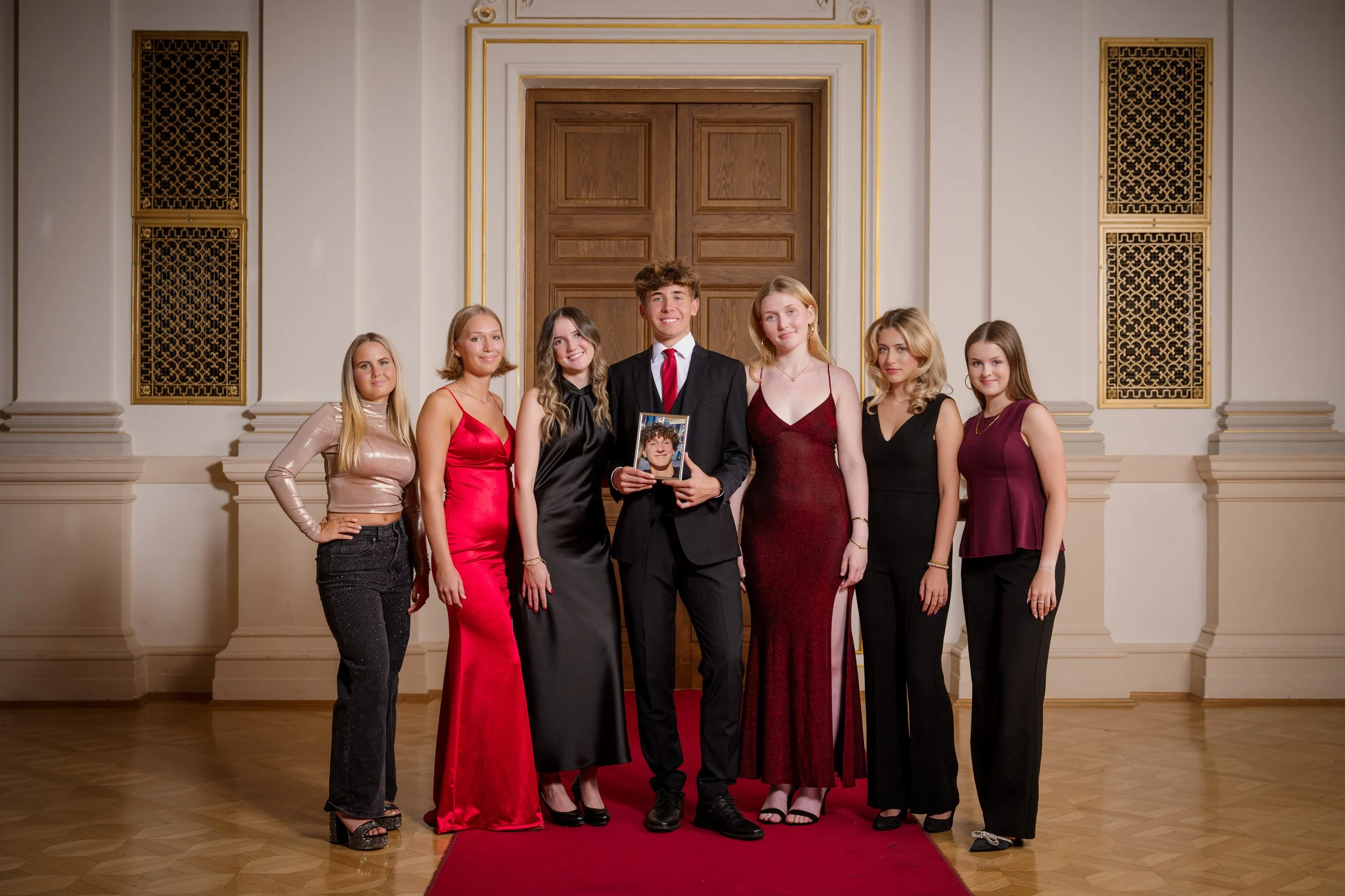 Eine Gruppe von acht jungen Menschen in formeller Kleidung posiert auf einem roten Teppich vor einer großen Holztür mit goldene Dekor. Sie scheinen an einem besonderen Anlass teilzunehmen.