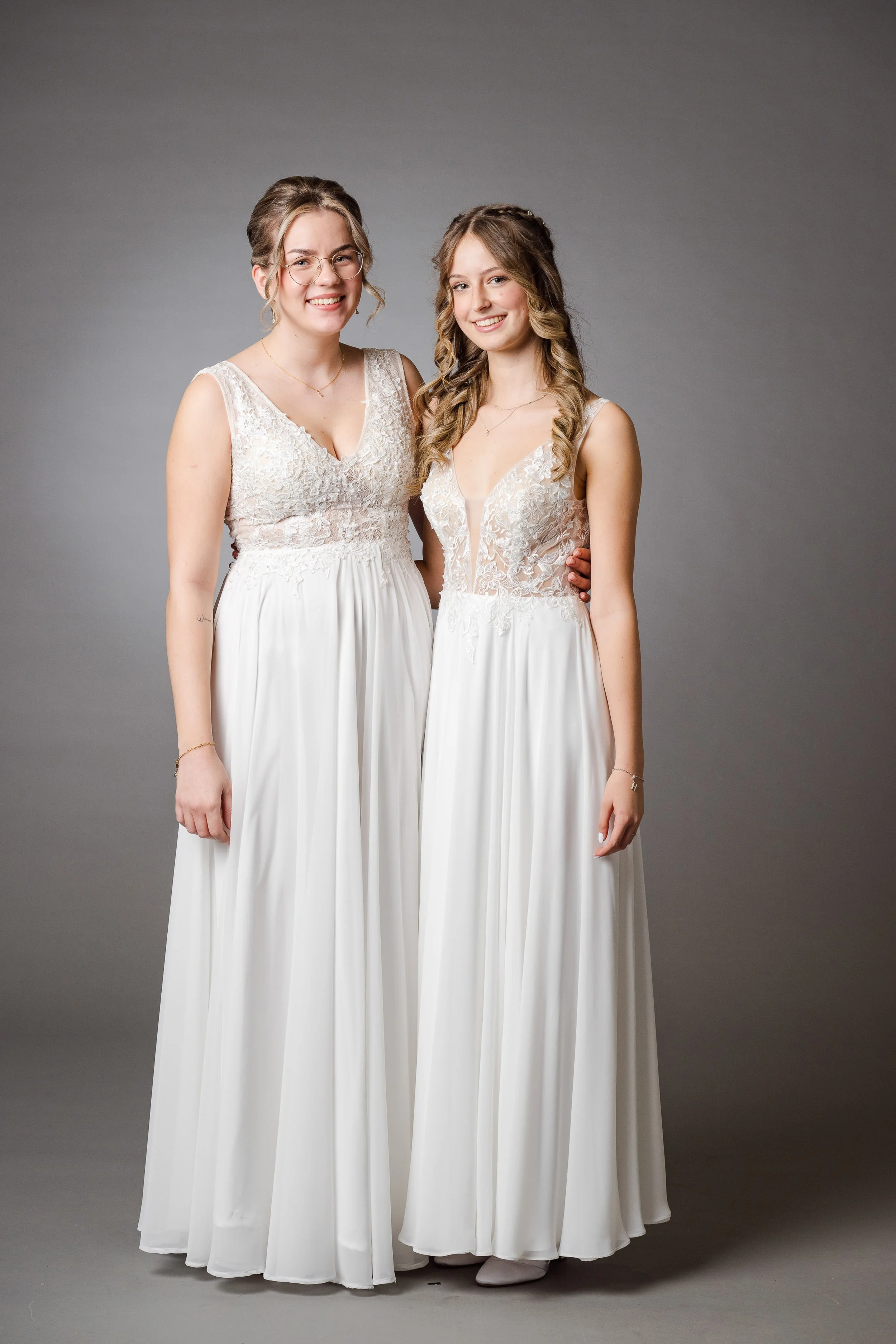 Zwei Frauen in eleganten weißen Brautkleidern posieren vor grauem Hintergrund.