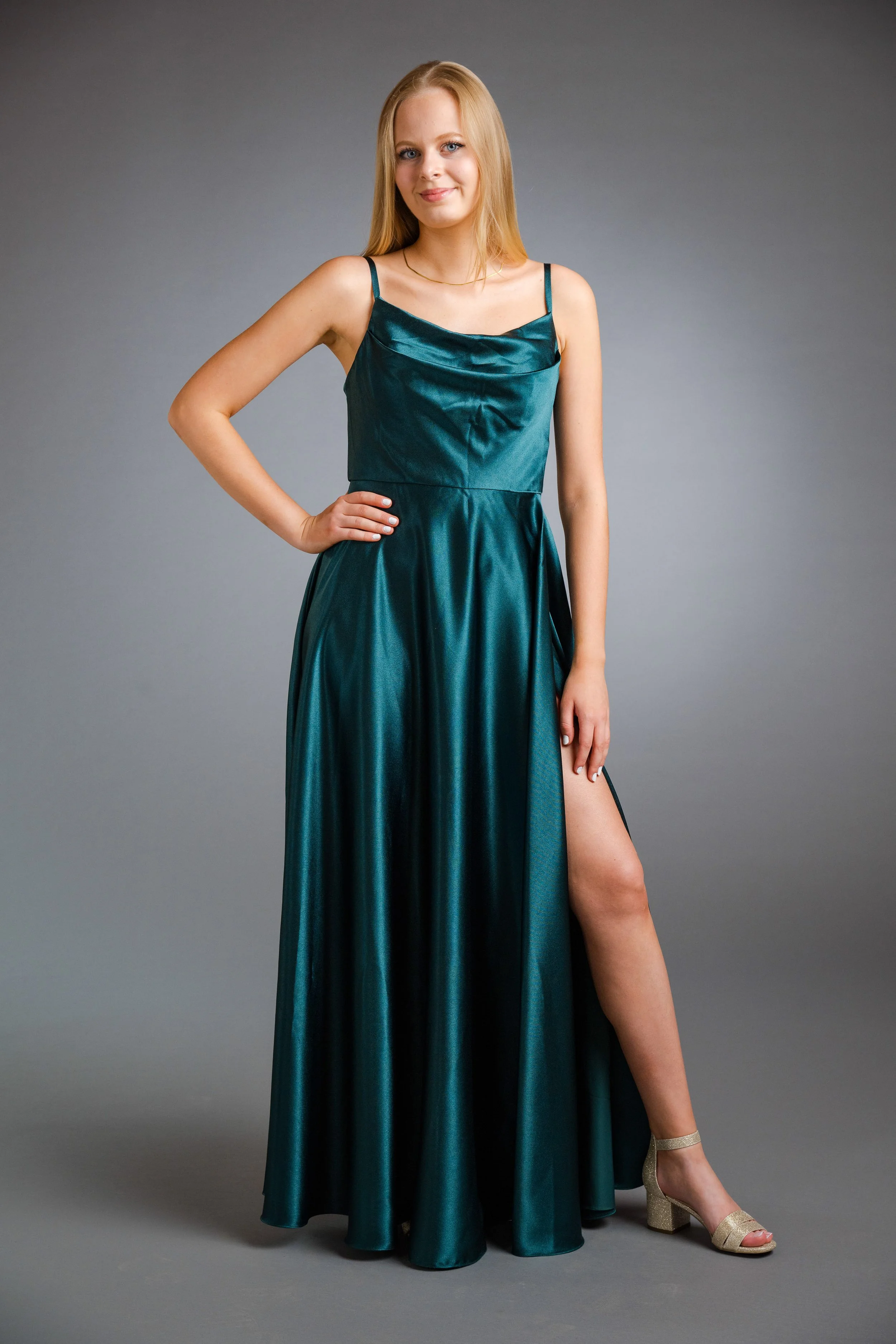Eine junge Frau in einem eleganten, smaragdgrünen Abendkleid mit Schlitz, die auf einem grauen Hintergrund posiert.