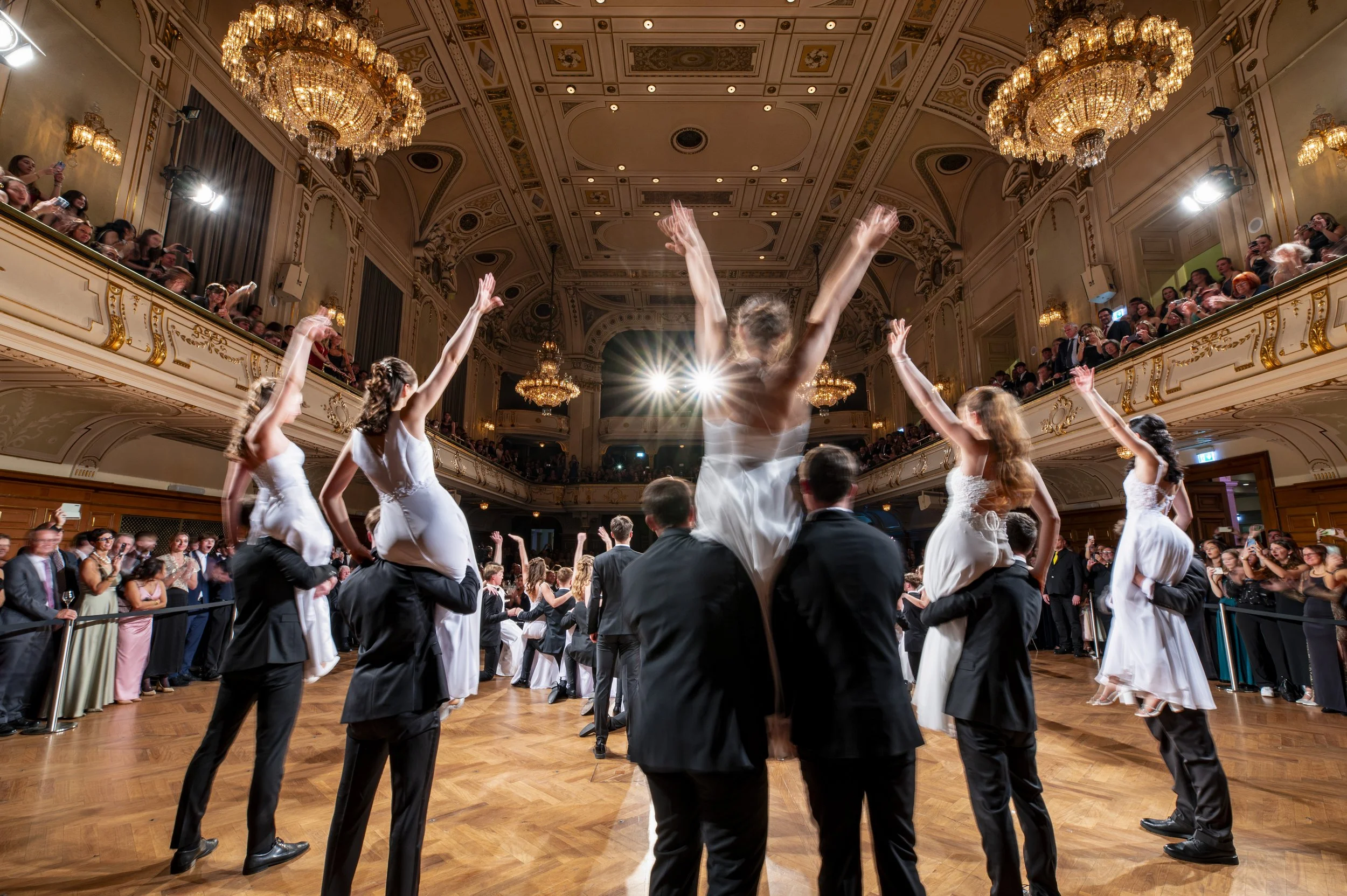Tanzender Hochzeitspaare in elegante Abendkleider und Anzüge, umgeben von applaudierendem Publikum in einem opulenten Ballsaal mit Kronleuchtern.
