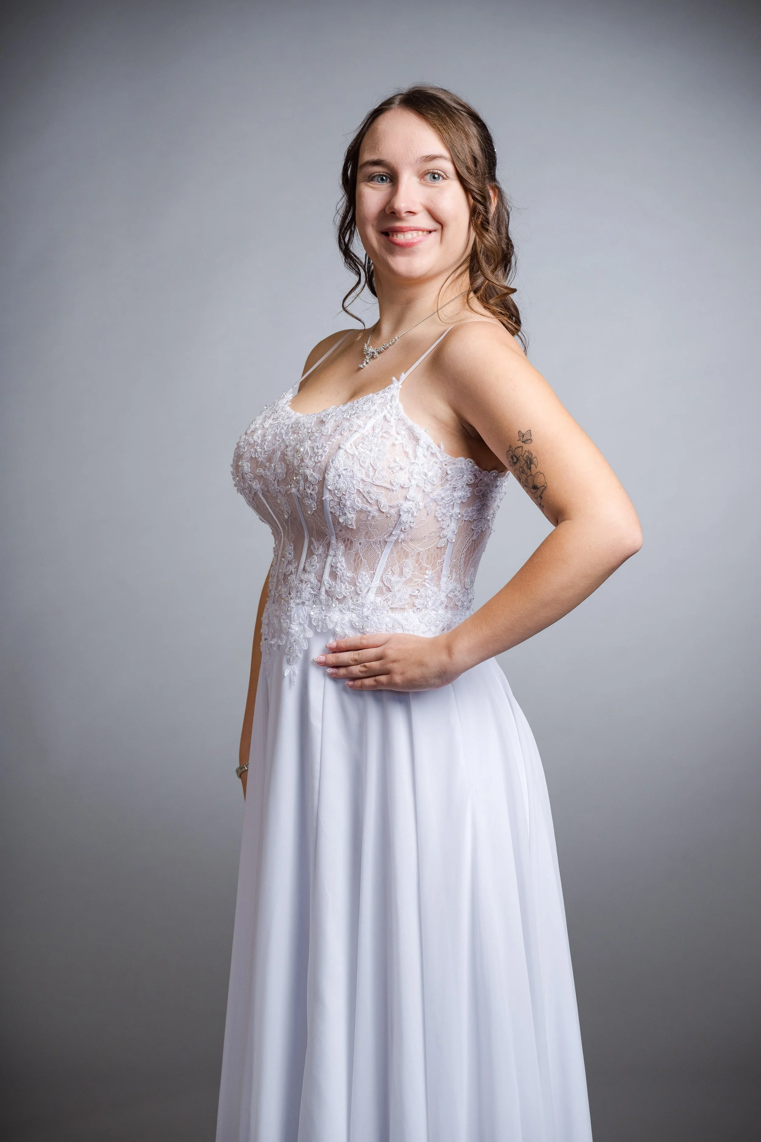 Junge Frau in Brautkleid, steht seitlich vor grauem Hintergrund, lächelt, trägt Schmuck, hat Tattoo am Arm.