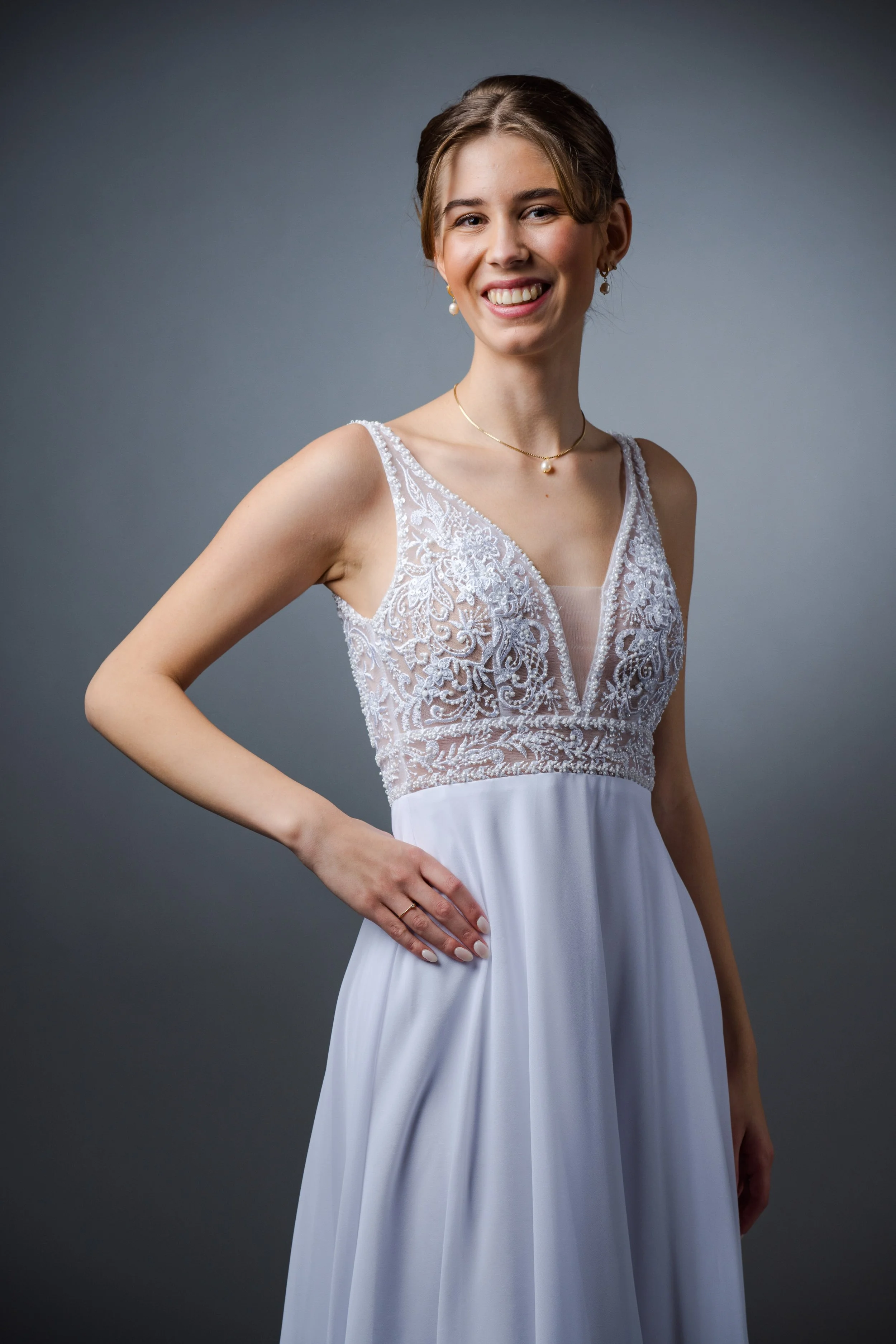 Junge Frau trägt ein weißes Brautkleid mit Spitzen- und Stickerei-Details, lächelt, vor grauem Hintergrund.