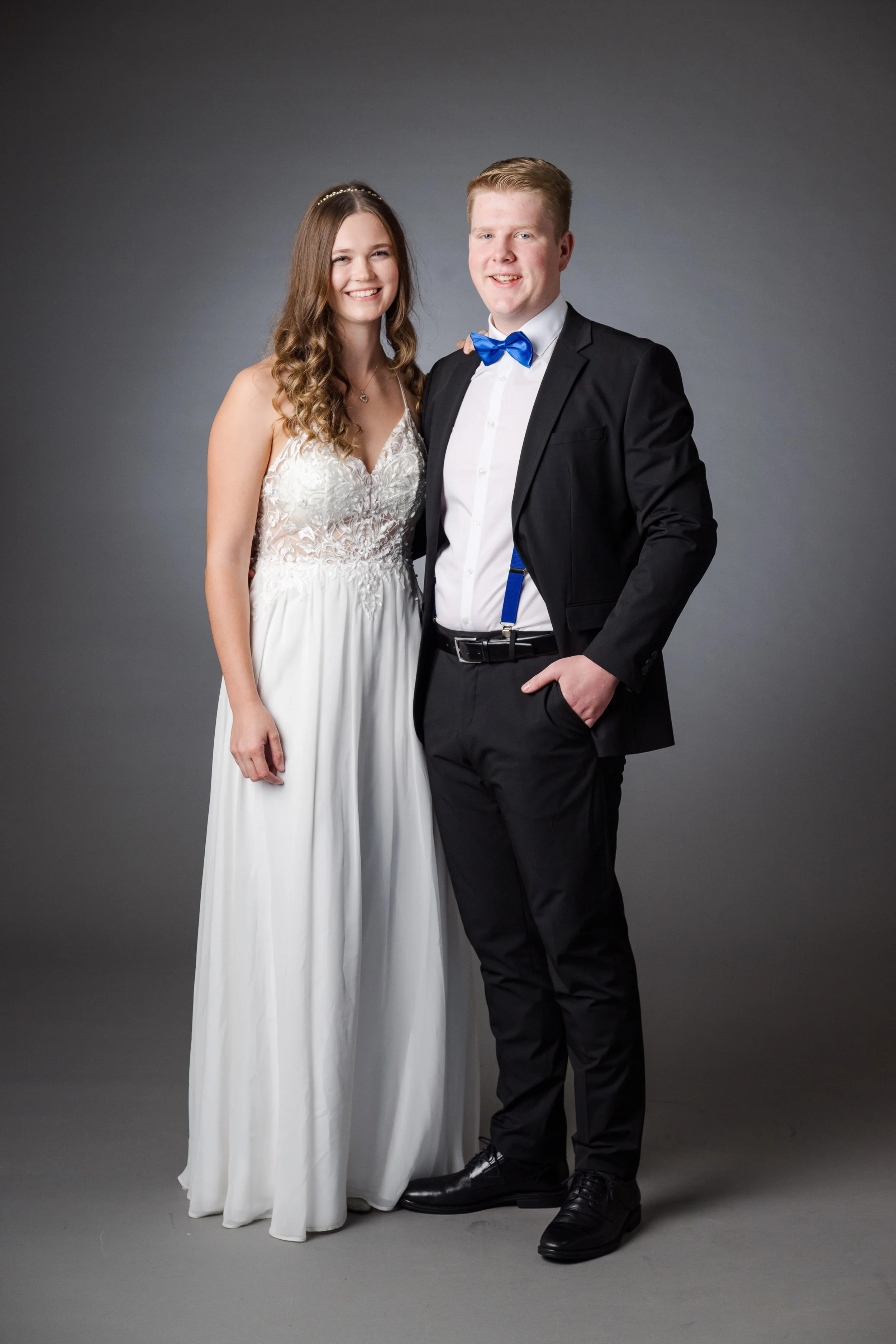 Ein junges Paar, das auf einer Fotoproduktion in formeller Kleidung posiert. Das Mädchen trägt ein weißes, langes Brautkleid mit Spaghettiträgern und das Junge trägt einen schwarzen Anzug mit blauer Fliege.