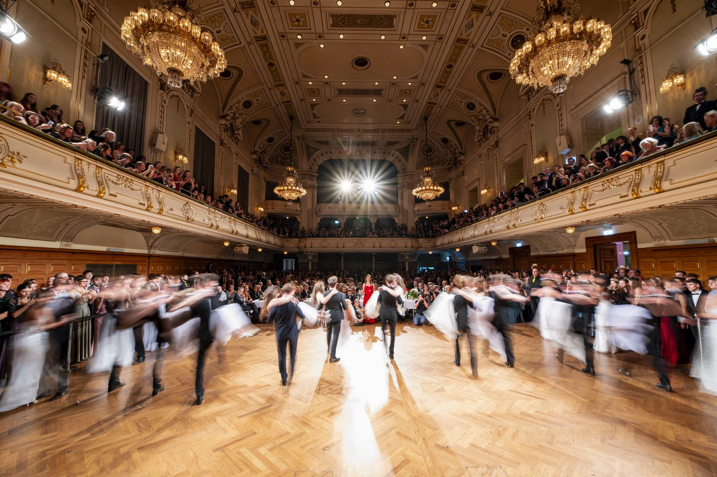 Ein festlich geschmückter Ballsaal mit einem Tanz-Event, auf der Bühne tanzen Paare im formellen Kleidung, während im Hintergrund viele Zuschauer sitzen, die den Tanz beobachten. Die Szene ist elegant, mit goldverzierten Wänden, Kronleuchtern und ein