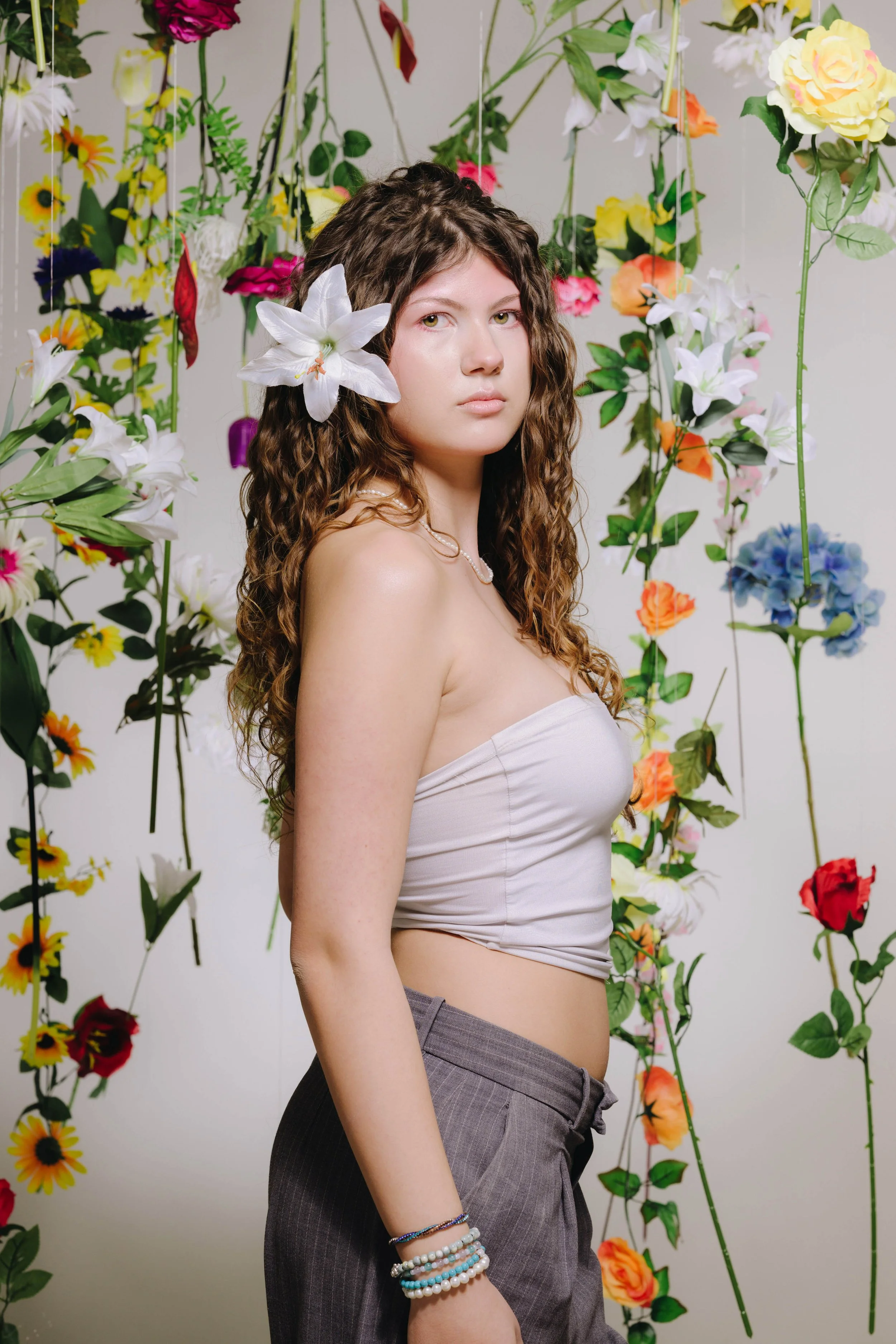 Junge Frau mit Locken, trägt ein weißes Top und graue Hosen, hat eine weiße Lilie im Haar, steht vor einer Wand mit hängenden, bunten Blumen.