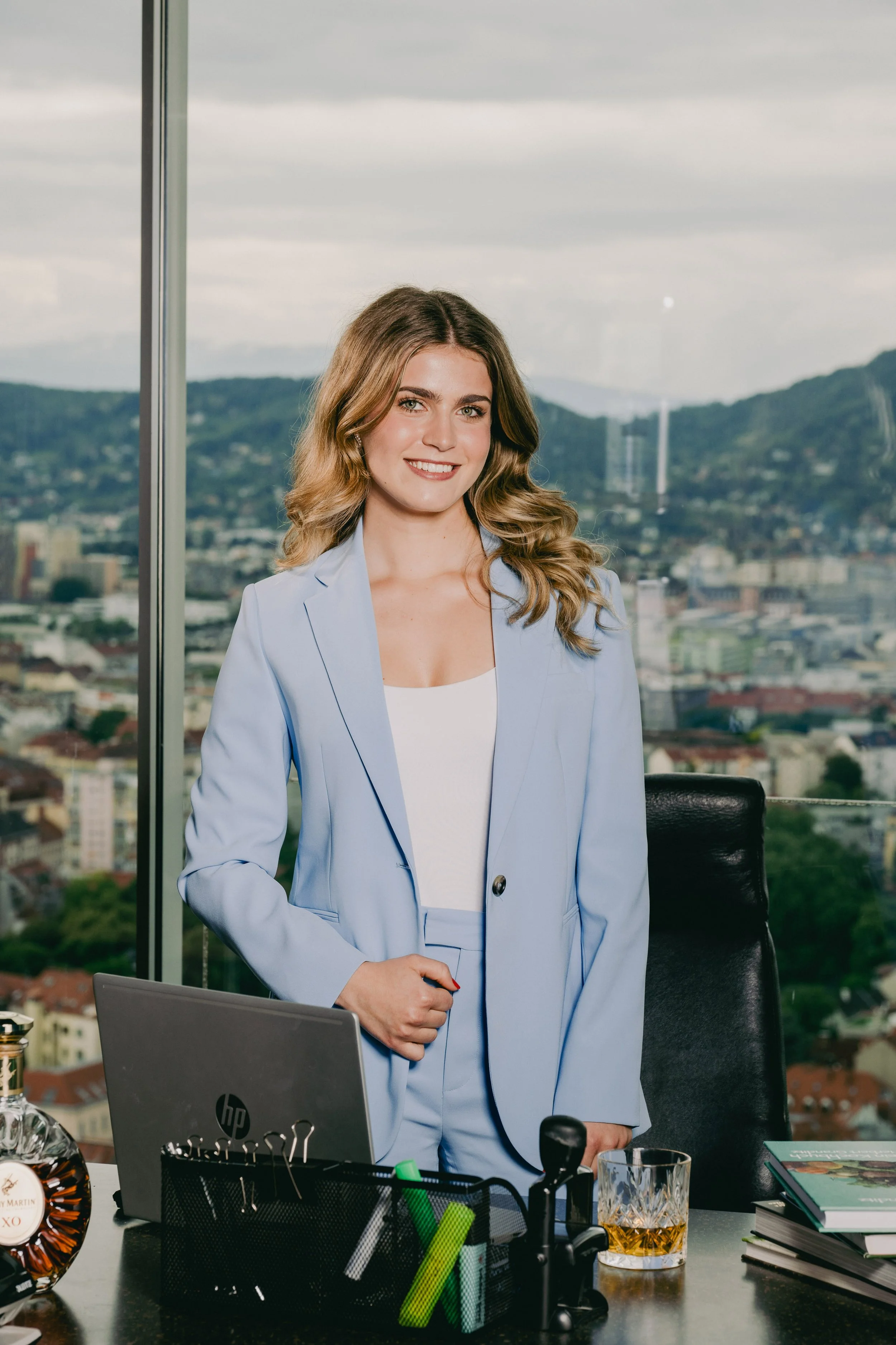 Eine Frau in einem hellblauen Business-Outfit steht in einem Büro vor einem Fenster mit Stadt- und Hügelblick. Sie lächelt und hat lang, gewellte, braune Haare.