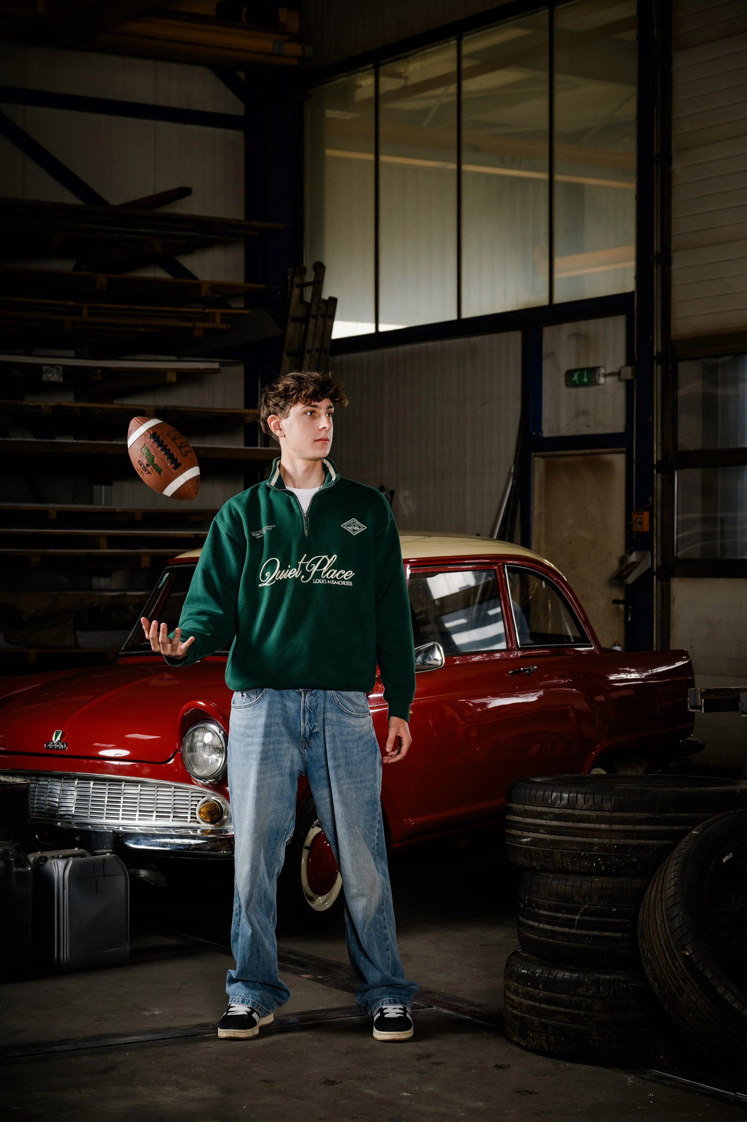 Junge wirft Football in einem Autohaus, hinter ihm ein rotes Auto, umgeben von Reifen und einem schwarzen Koffer, im Hintergrund Regale und ein Lader.