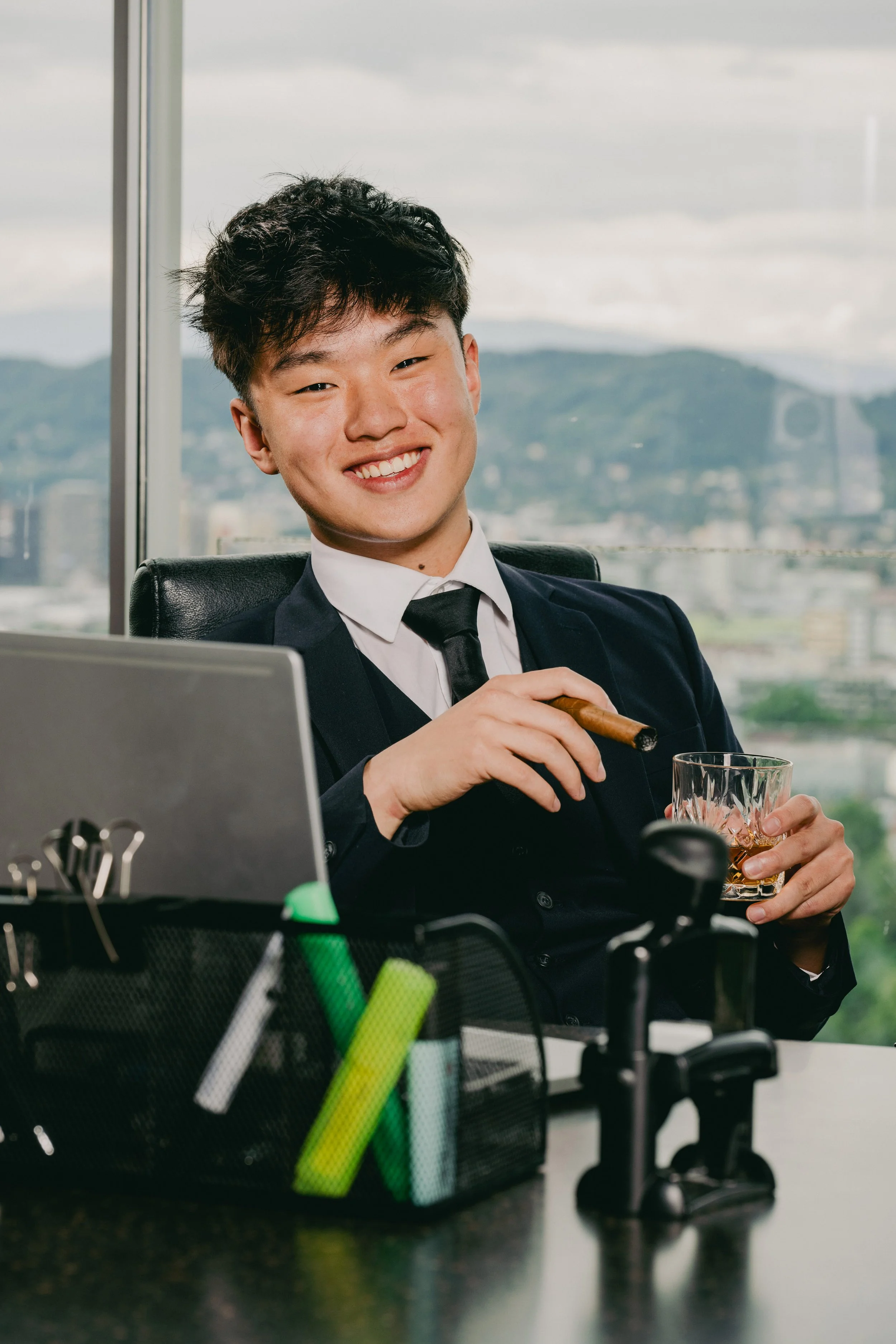 Ein junger Mann im Anzug sitzt an einem Schreibtisch, lächelt und hält eine Zigarre sowie ein Glas Whiskey, im Hintergrund eine Stadt- und Berglandschaft bei bewölktem Himmel.