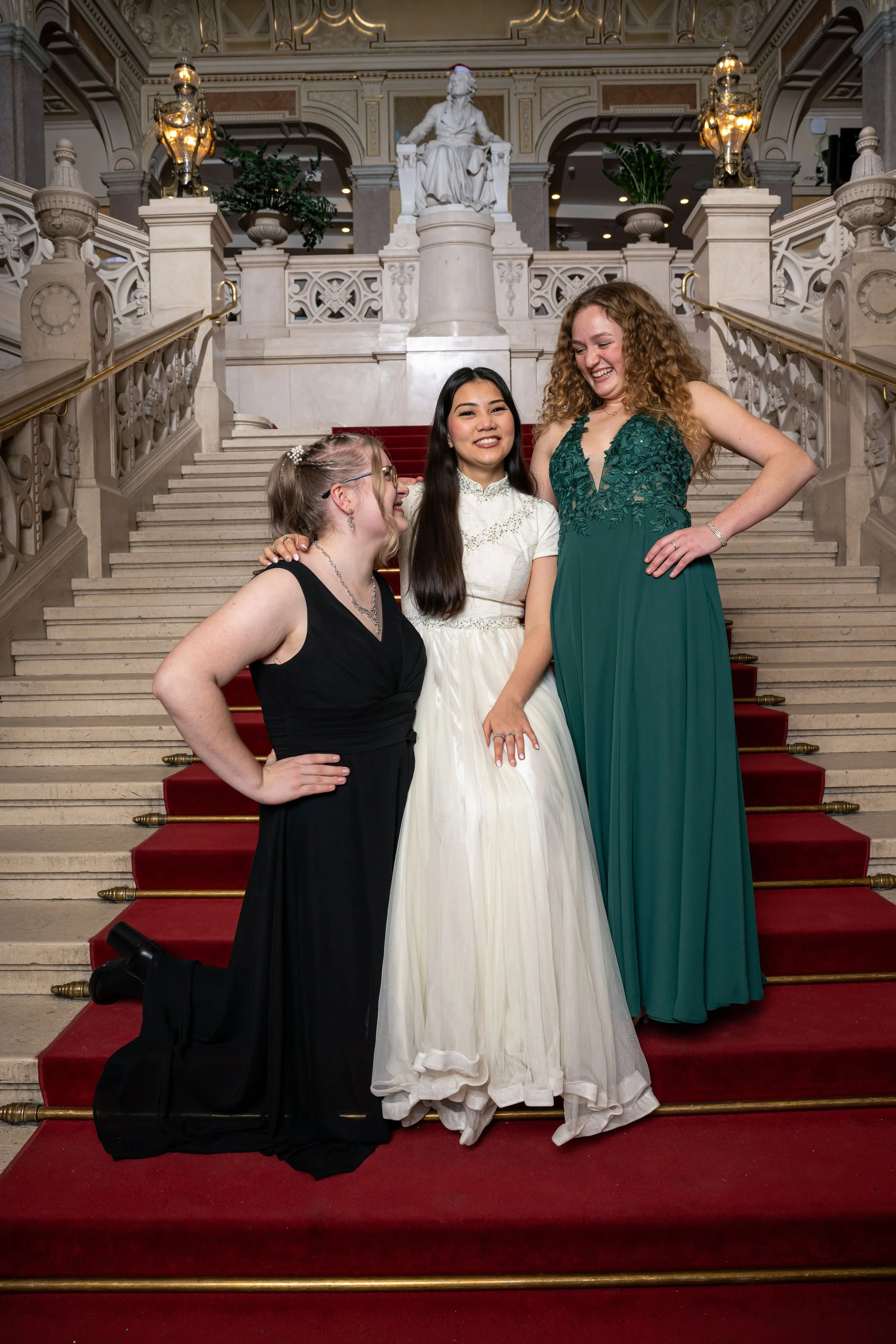 Vier Frauen in eleganten Abendkleidern posieren fröhlich auf einer Treppe in einem prunkvollen, historischen Gebäude mit Statuen und goldenen Leuchtern im Hintergrund.