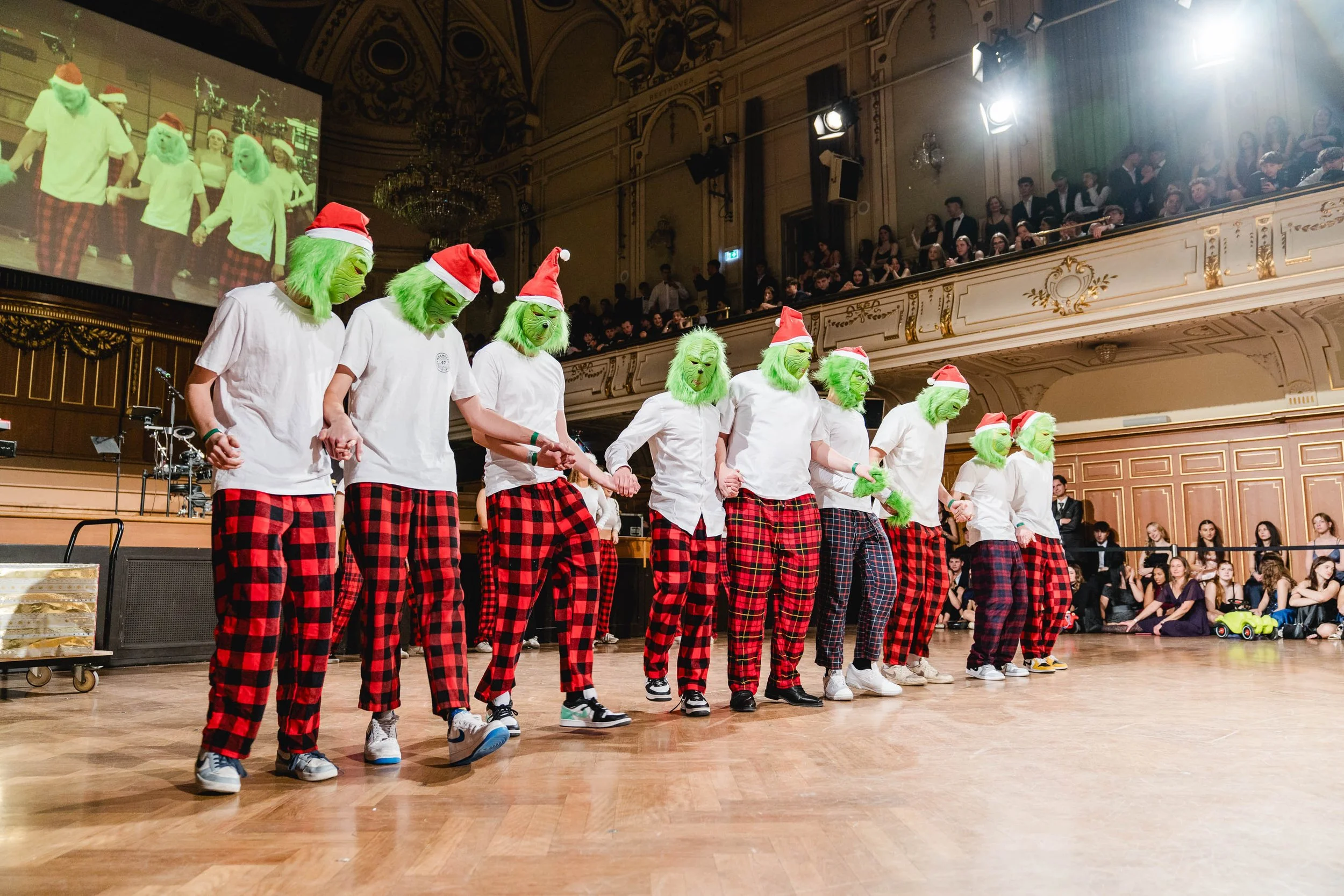 Gruppe von Menschen in Grinch-Masken, weißen T-Shirts und roten Schottenkaro-Hosen, die Hand in Hand in einem Ballsaal tanzen. Die Masken mit grüner Haut und Weihnachtsmützen. Zuschauer sitzen im Hintergrund auf der Galerie.