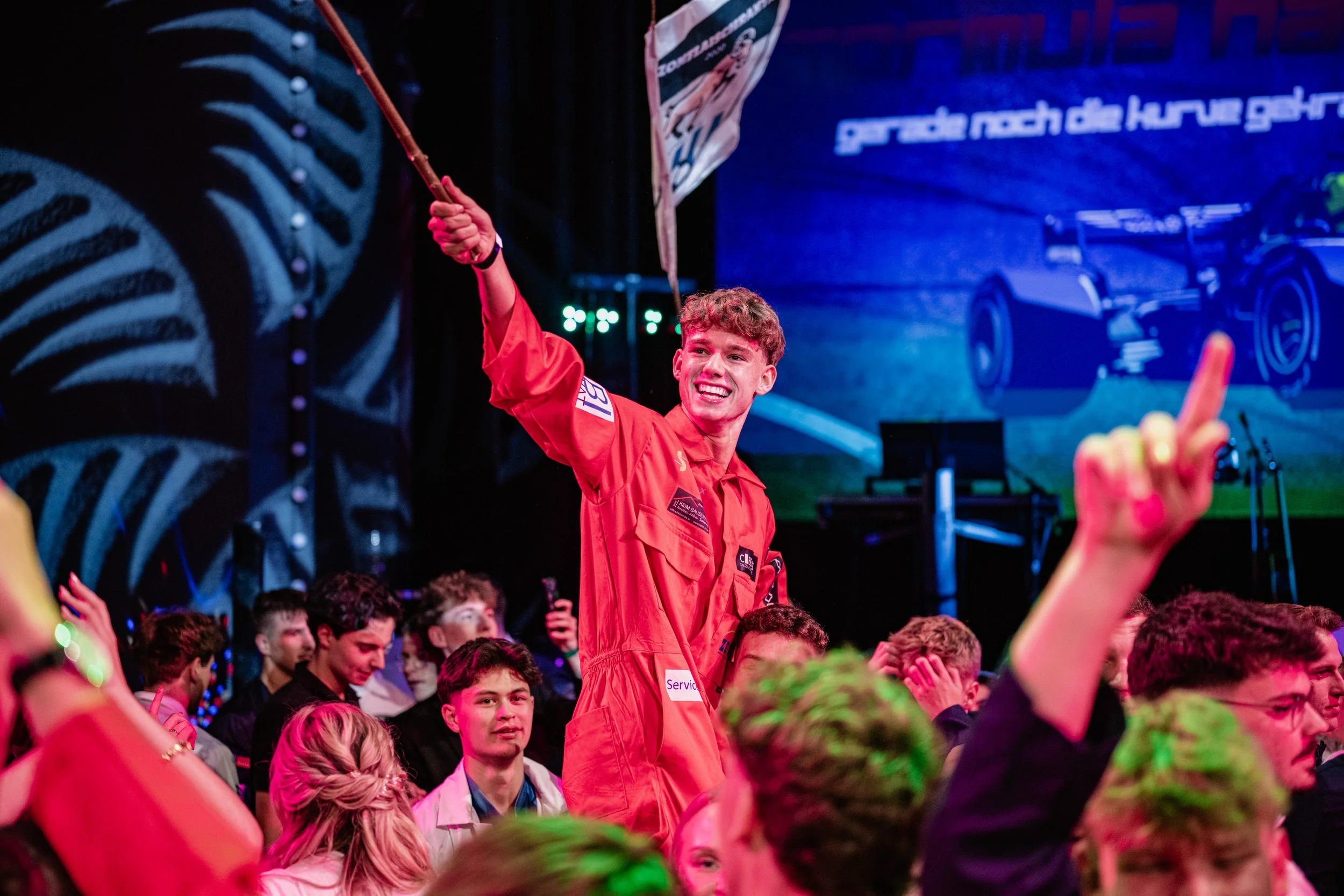 Junger Mann in rotem Race-Overall auf einer Veranstaltung, auf einem Podium, winkt mit einem Flaggenstab, lächelt. Im Hintergrund eine große Bildschirmprojektion eines Formel-1-Autos.