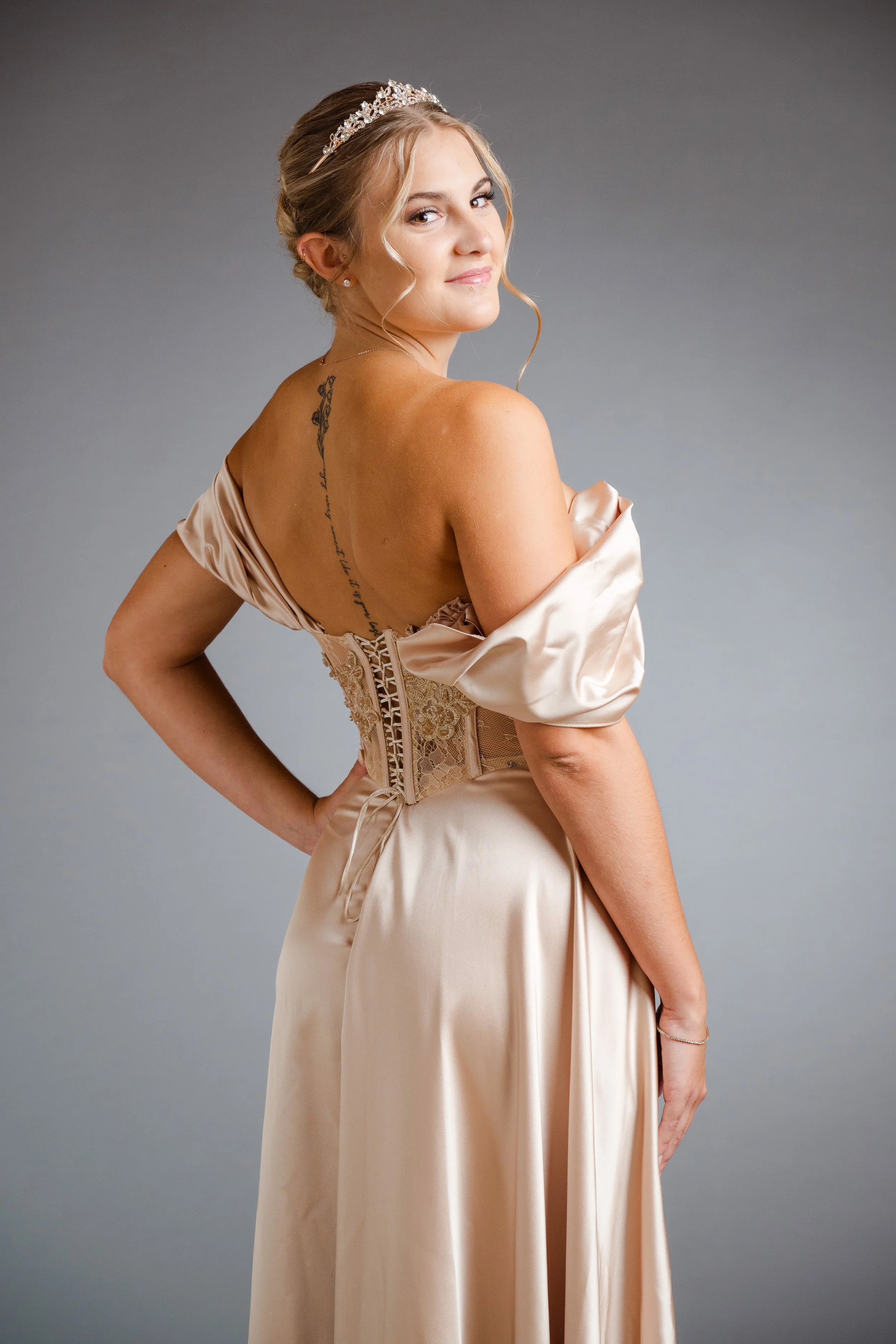 Eine Frau in einem eleganten, cremefarbenen Brautkleid mit Schultern und einem leichten Lächeln, trägt eine Tiara und hat eine Tätowierung auf dem Rücken.