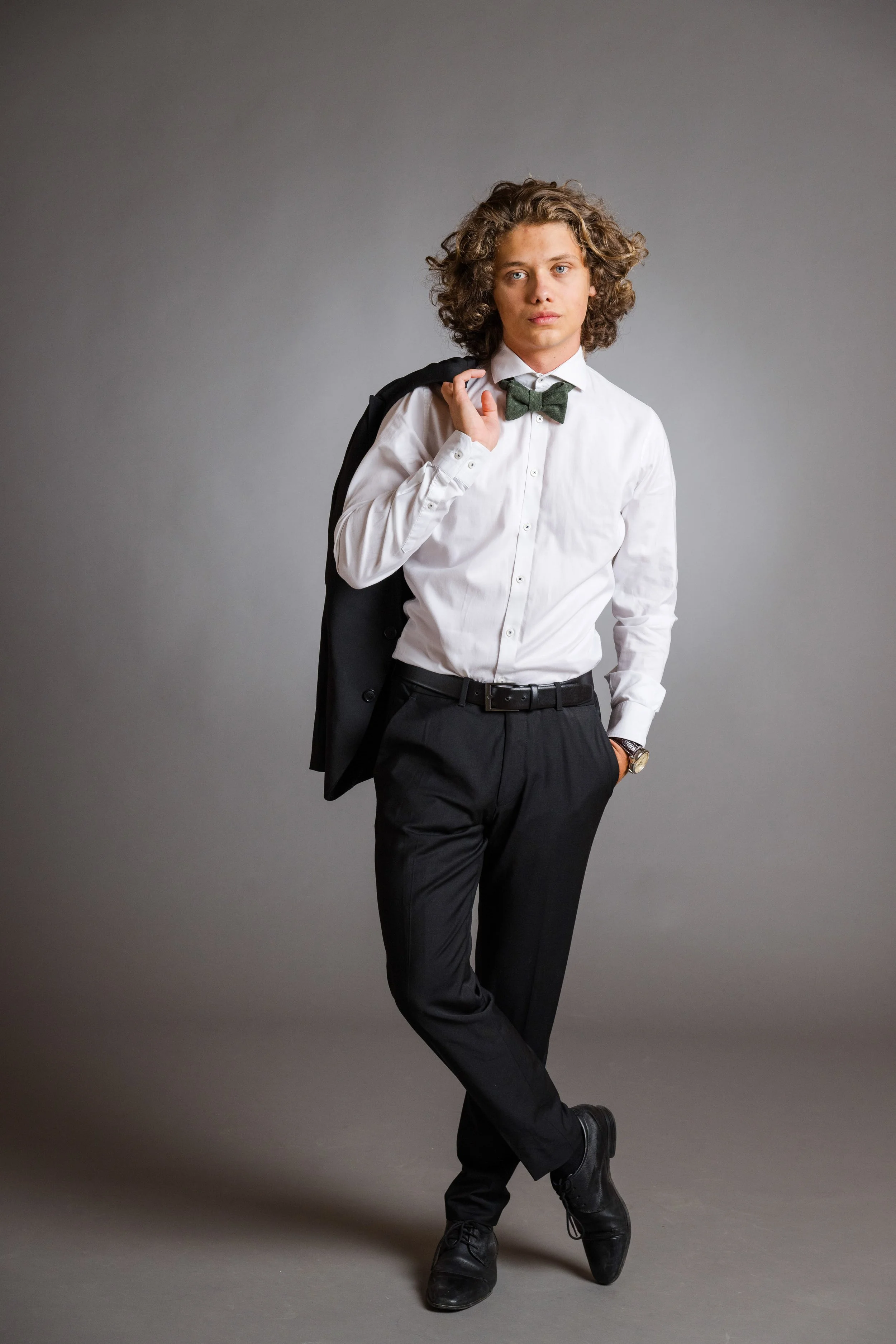 Junge männliche Person mit lockigem Haar trägt weiße Hemd, grüne Fliege, schwarze Hose, schwarze Schuhe und hält schwarzen Blazer über der Schulter. Neutraler Hintergrund.