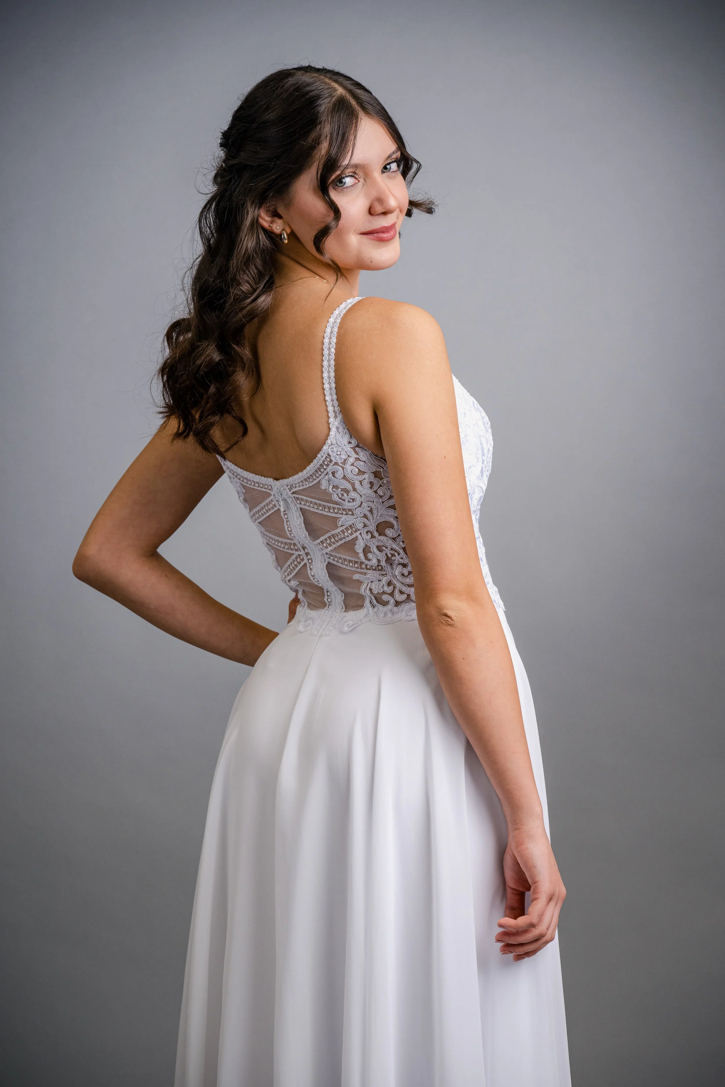 Eine junge Frau in einem weißen Brautkleid mit Spitzenärmel posiert vor einem grauen Hintergrund.