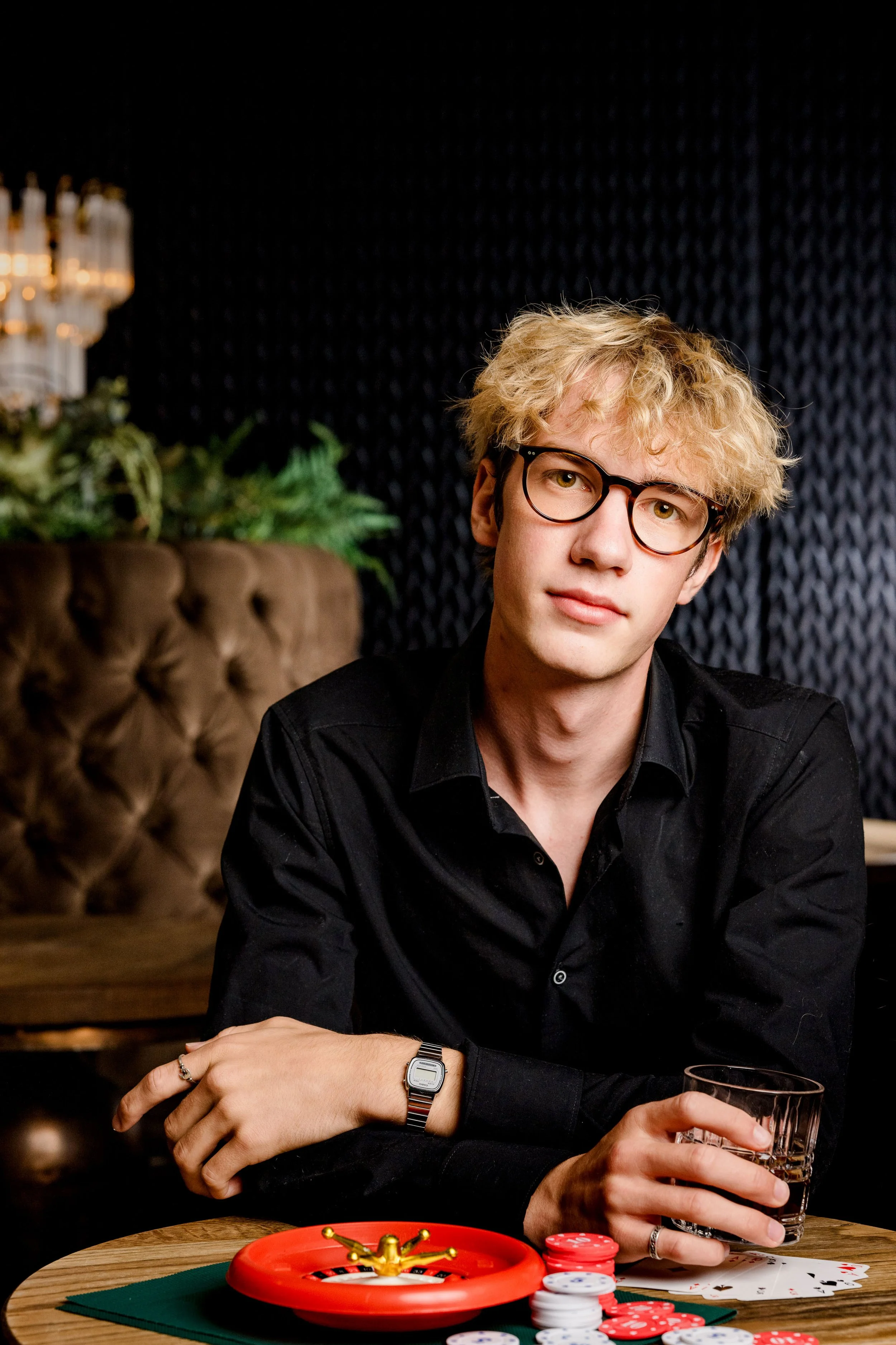 Ein junger Mann mit Brille und blondem, lockigem Haar sitzt an einem Pokertisch in einem stilvollen Raum, hält ein Glas in der Hand, es liegen Spielchips und Karten vor ihm auf dem Tisch.