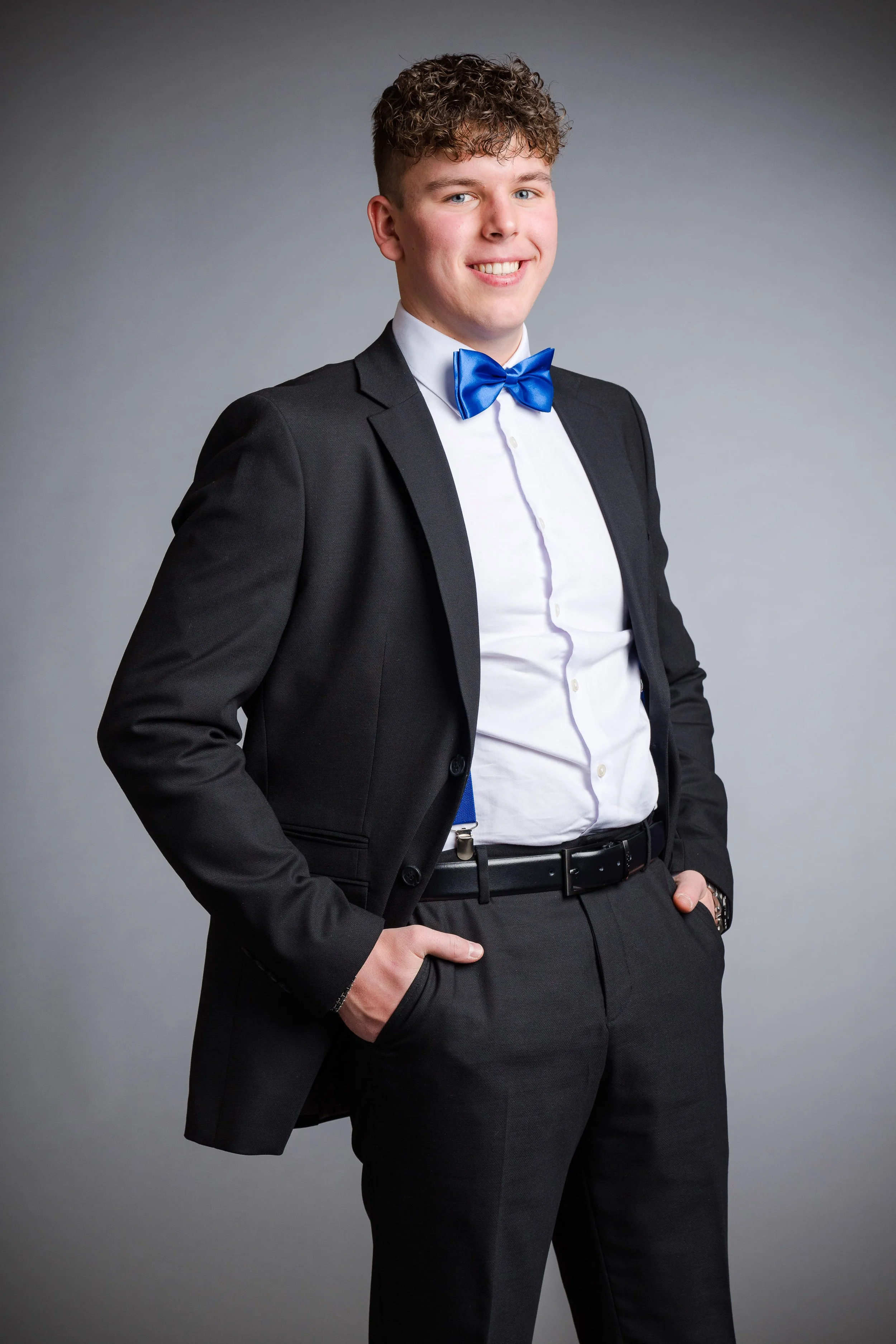 Junge Mann trägt formelle Kleidung mit schwarzem Anzug, weißem Hemd und blauer Fliege, lächelnd vor grauem Hintergrund.