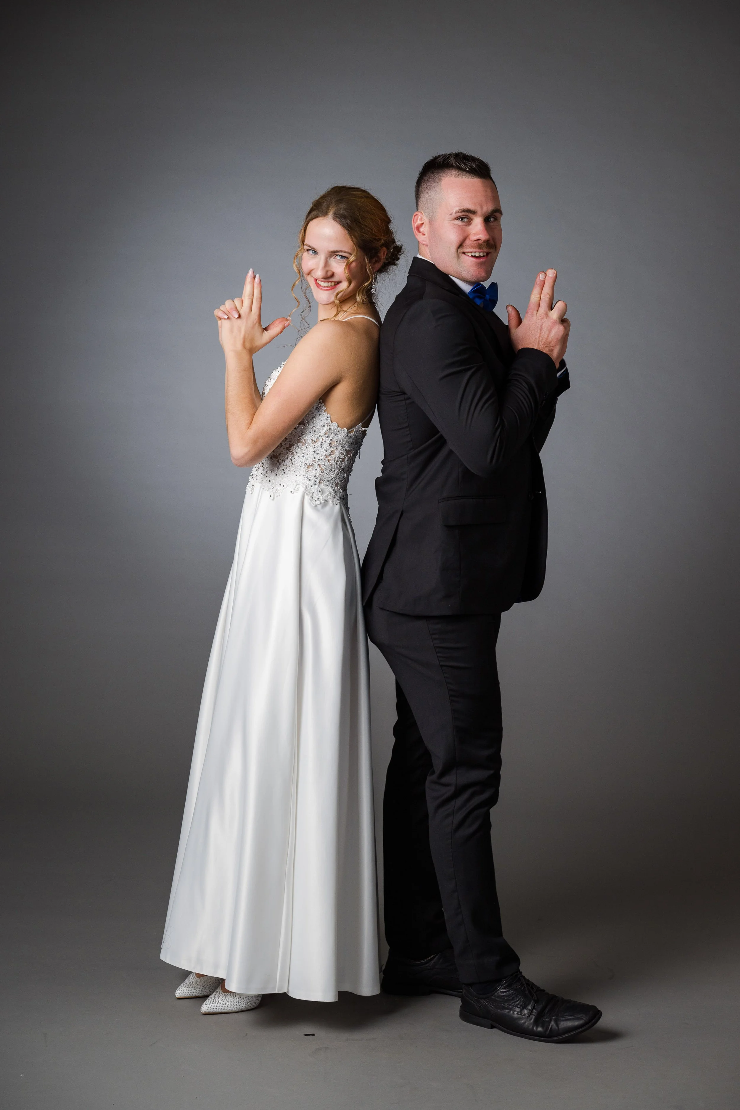 Ein Pärchen in Hochzeitskleidung, Rücken an Rücken, lächelnd, vor grauem Hintergrund.