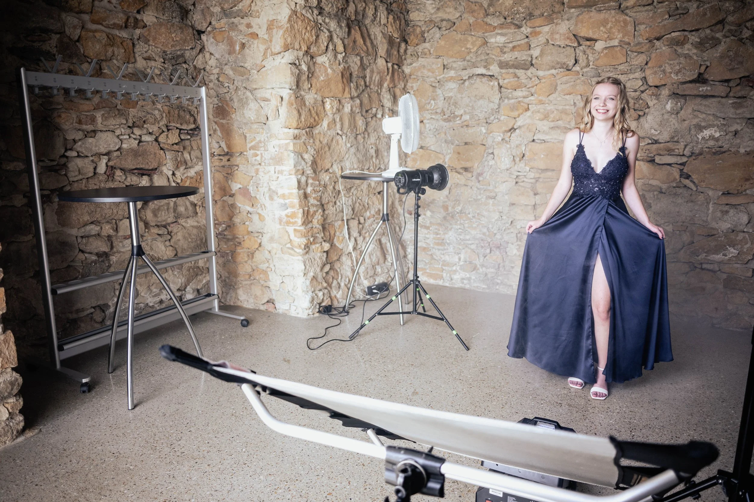 Eine lachende Frau in einem eleganten schwarzen Abendkleid steht vor einer Steinwand im Fotostudio und posiert.