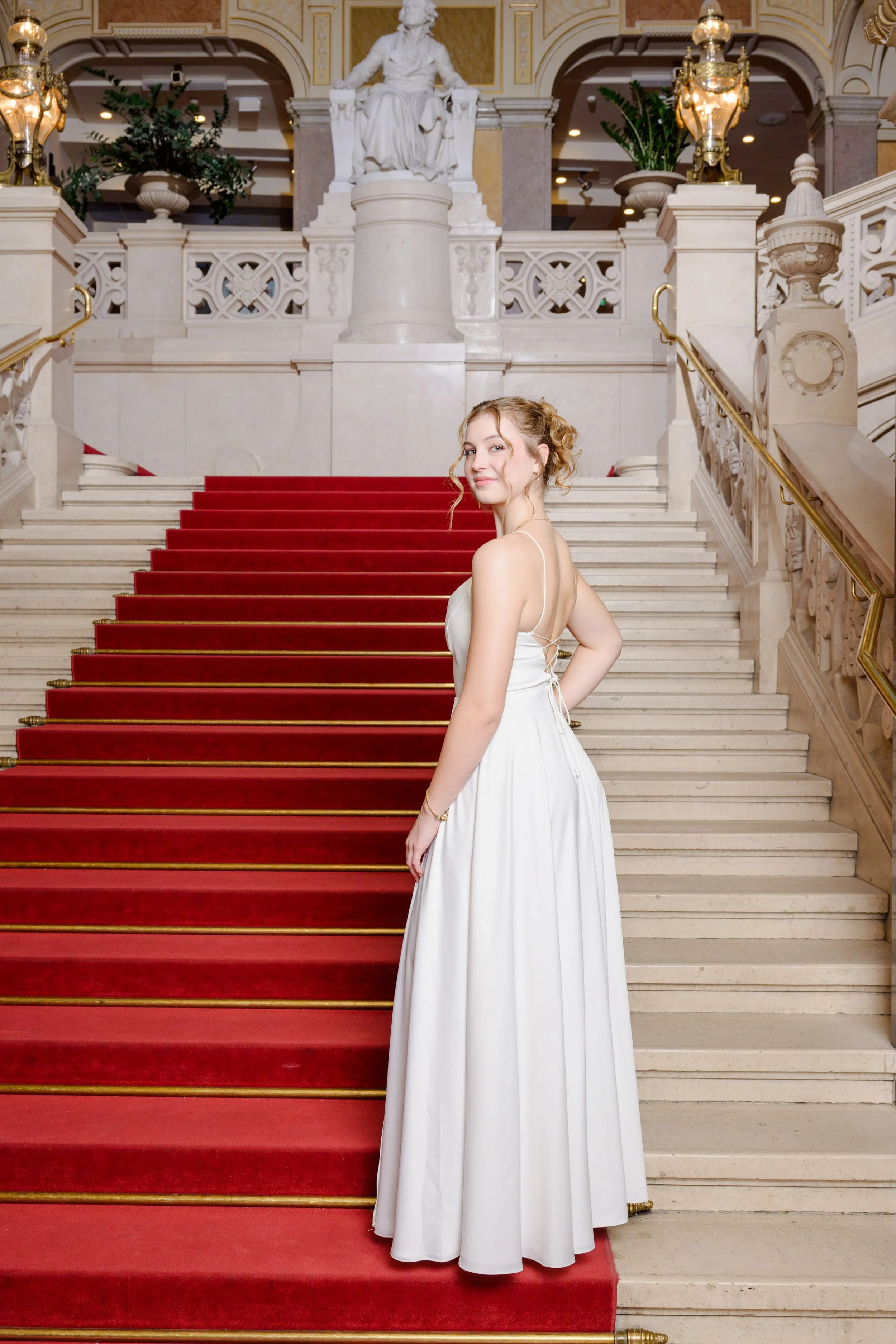 Junge Frau in einem weißen Kleid steht auf einer roten Treppe in einem prunkvollen, historischen Gebäude mit Marmors überdachter Treppe, Goldverzierung, einer weißen Statue im Hintergrund und Lampen an den Balustraden.