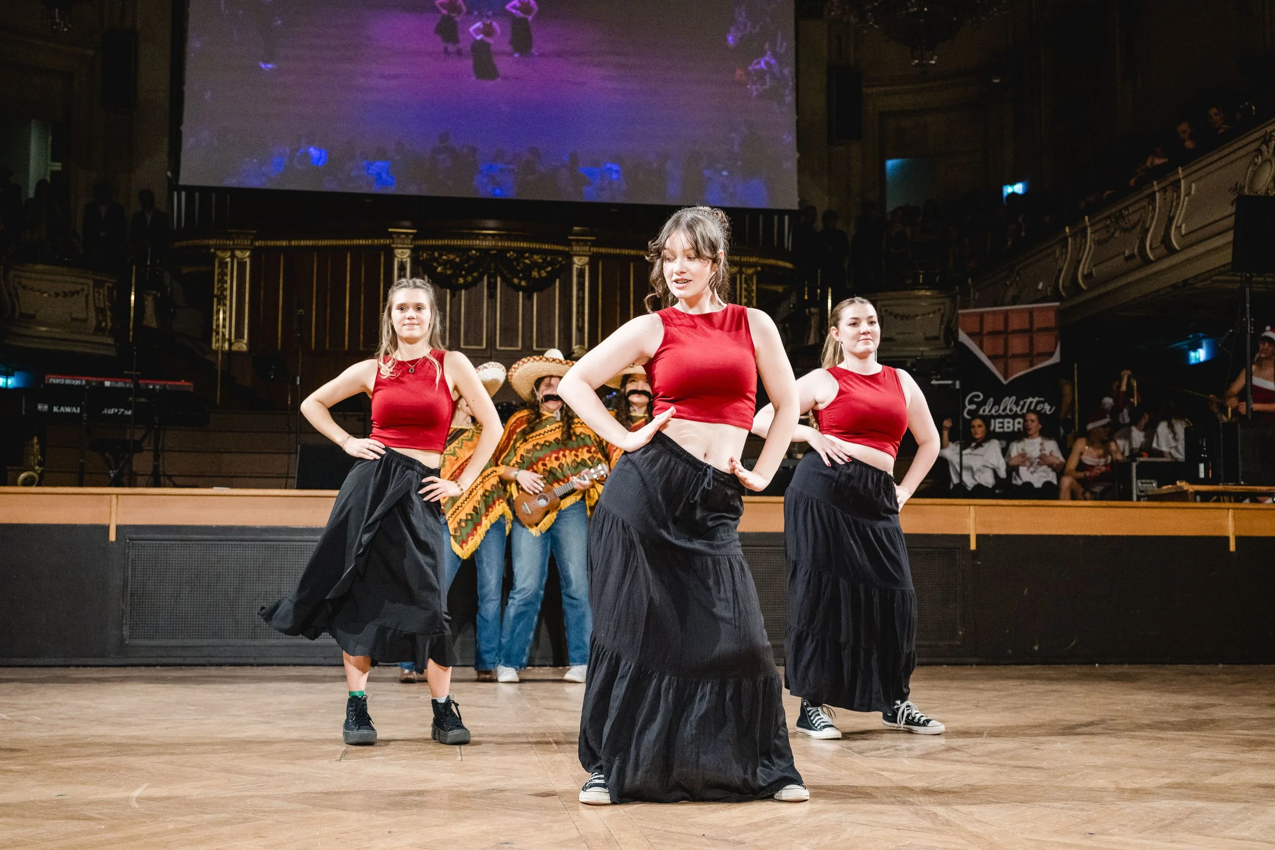 Drei junge Frauen tanzen auf einer Bühne, tragen rote Tops und schwarze Röcke, im Hintergrund sind Musiker mit Gitarren, und eine große Leinwand zeigt ihre Performance.