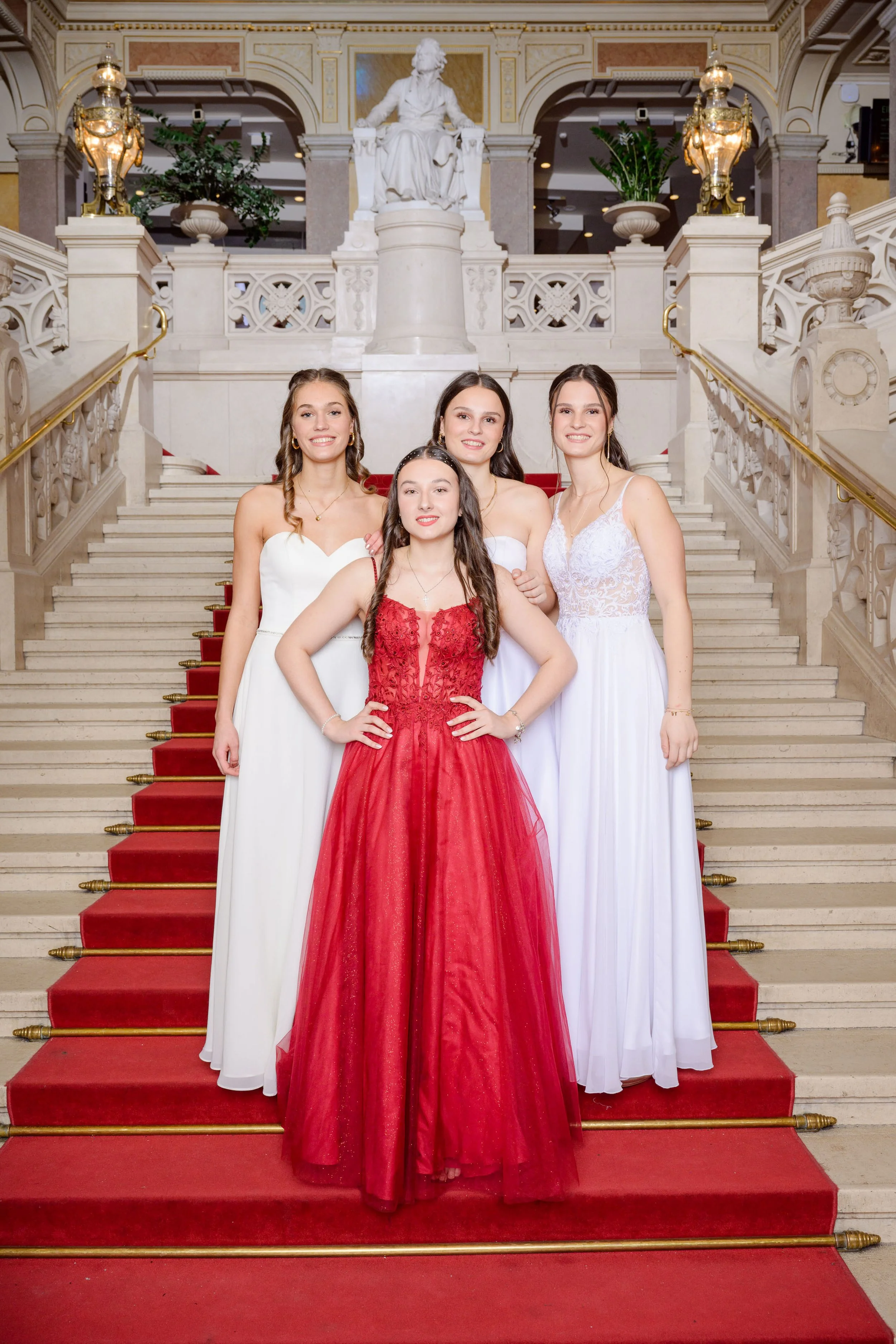 Fünf junge Frauen in eleganten Abendkleidern stehen auf einer Treppe vor einer Statue in einem prunkvollen Palastsaal.