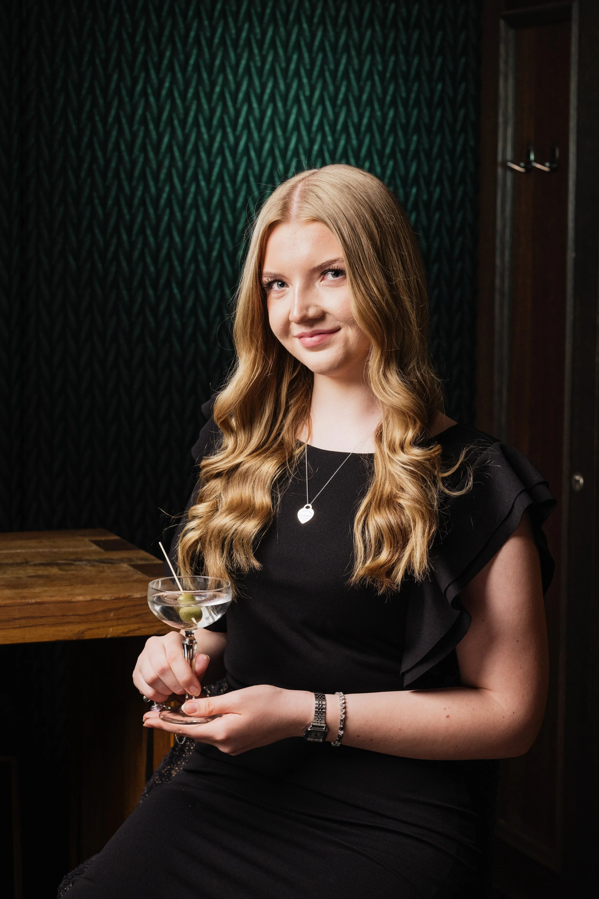 Junge Frau mit lang, gewelltem Haar, trägt Schwarz und hält einen Cocktail mit grünen Oliven, sitzt in einer Bar oder Lounge.