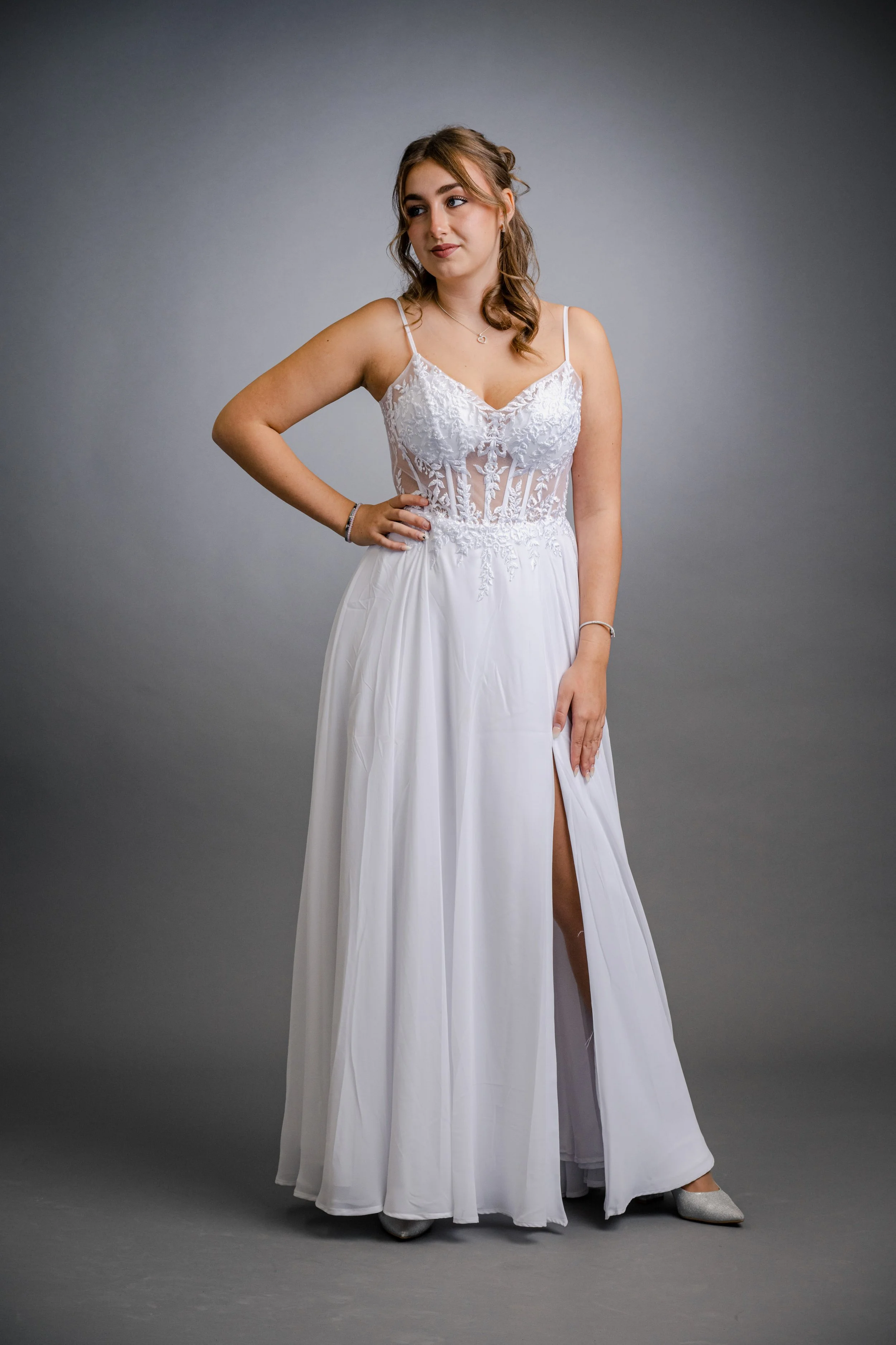 Junge Frau trägt ein weißes Hochzeitskleid mit Spitzen-Details, steht vor grauem Hintergrund, posiert mit Hand in der Taille, Blick seitlich.