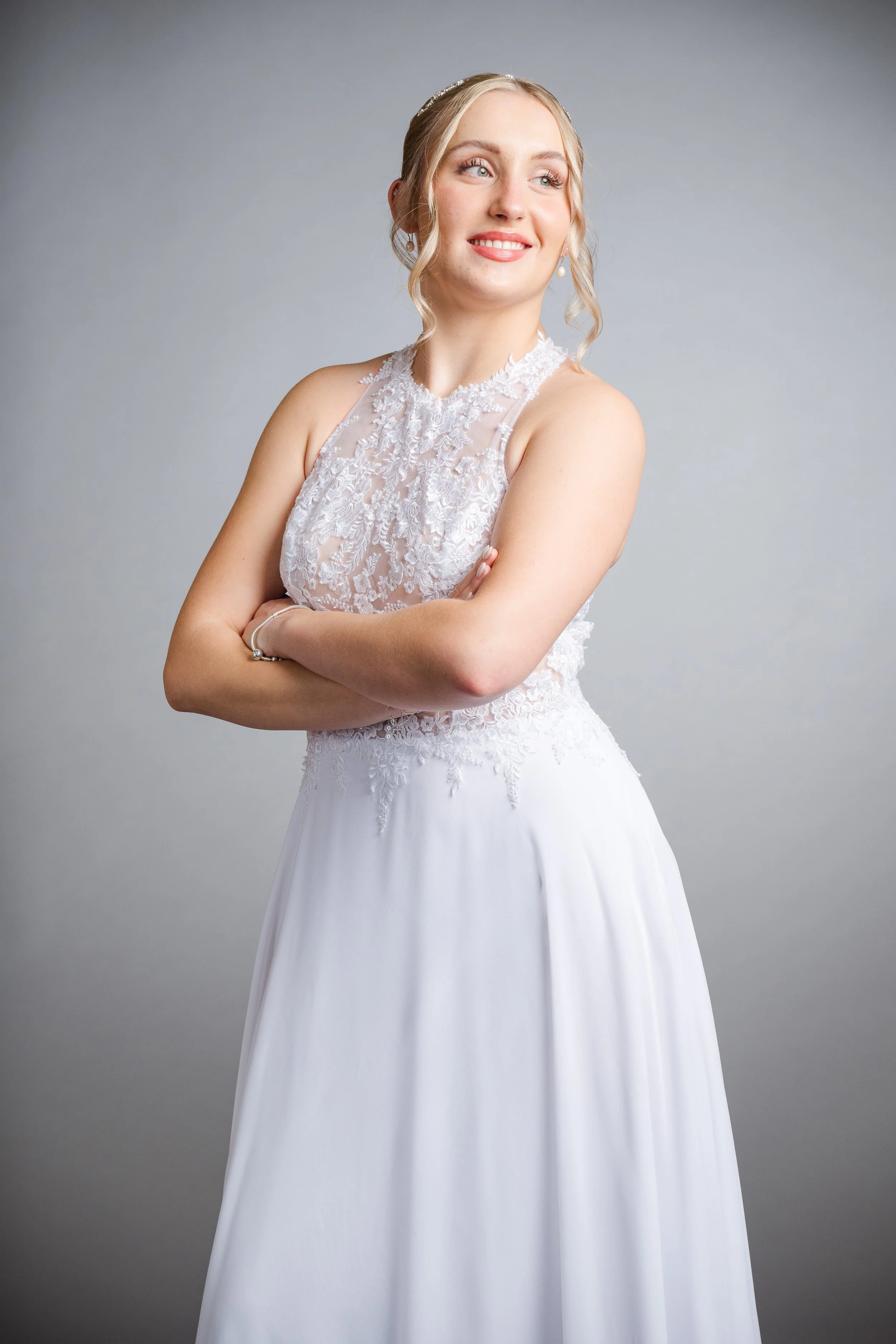 Eine junge Frau trägt ein weißes Hochzeitskleid mit Spitze und posiert vor einem grauen Hintergrund.