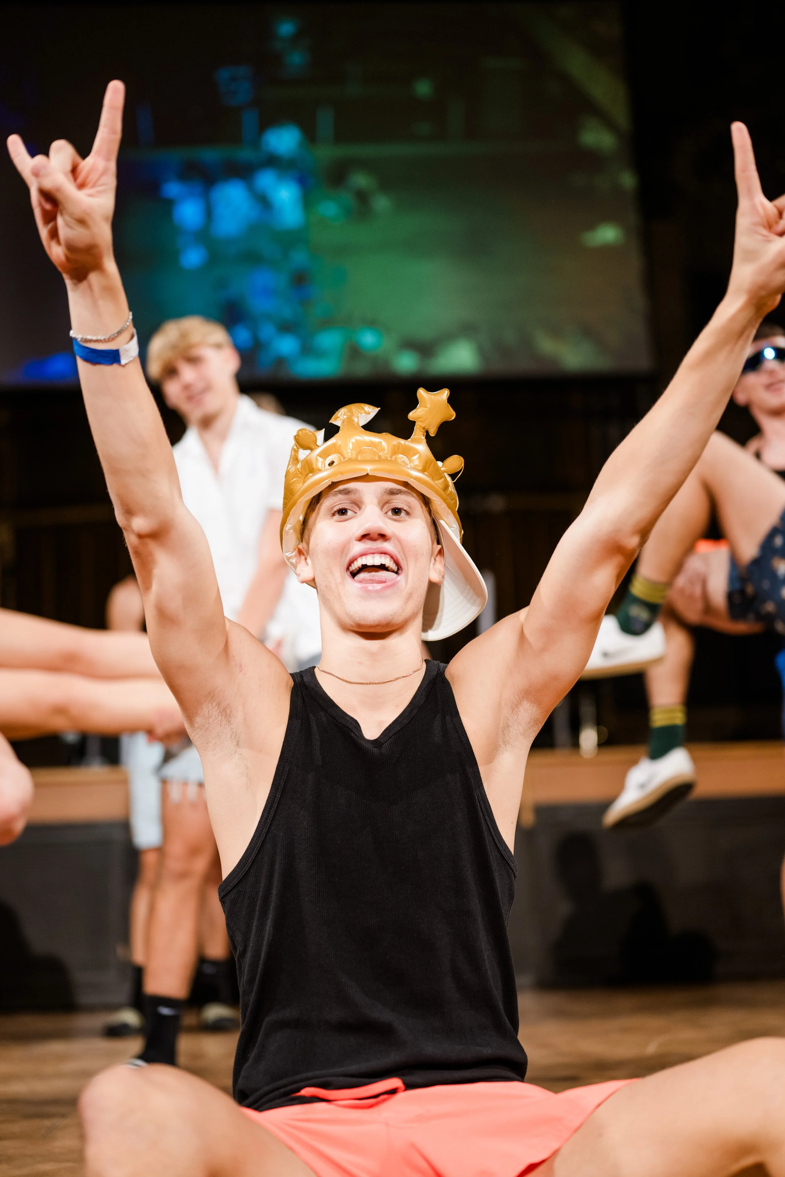 Junge mit goldener Partyhut, schwarzem Tanktop und roten Shorts, sitzt auf dem Boden, hebt die Arme in die Luft und lächelt bei einer Feier