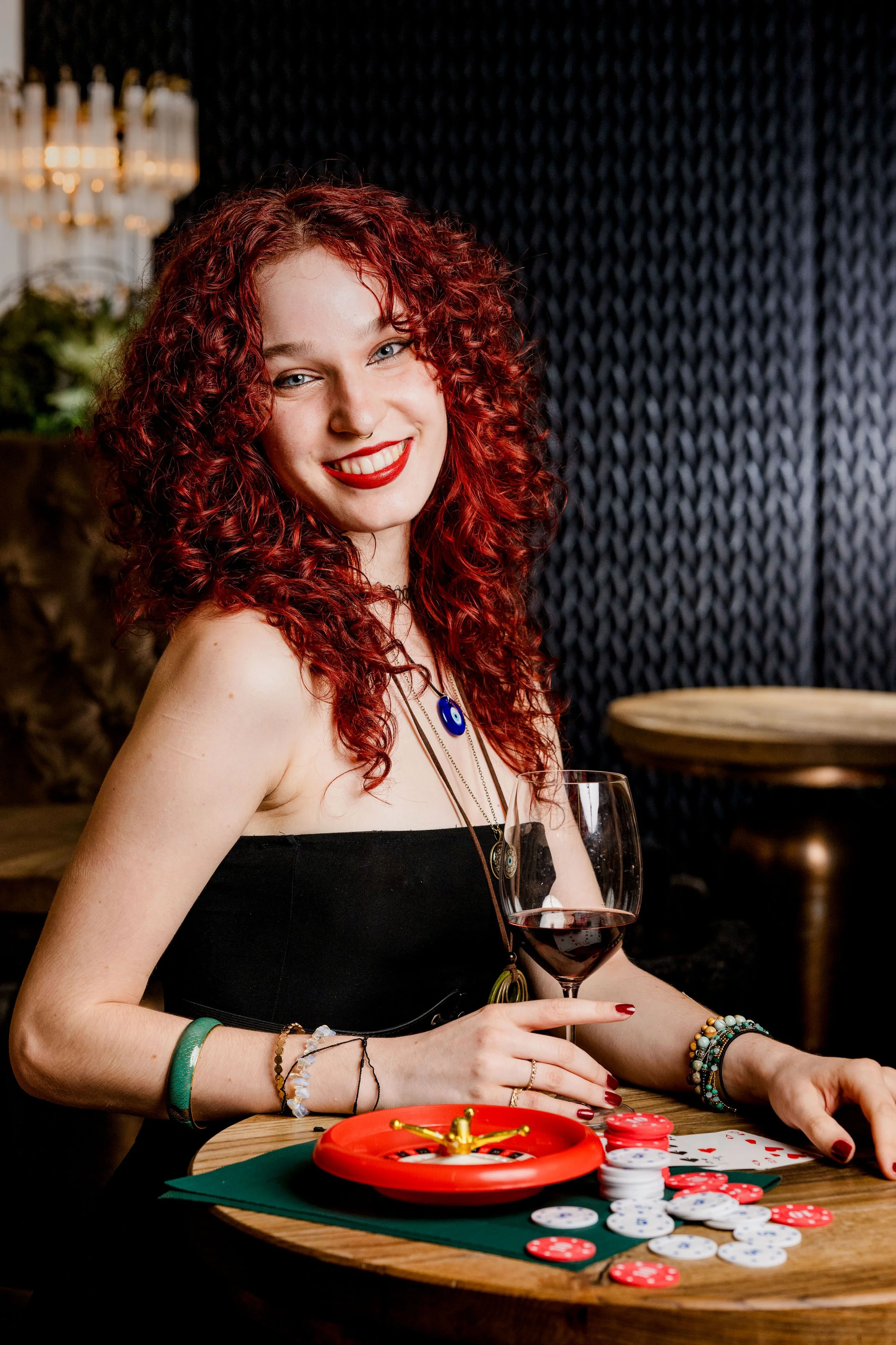 Eine lachende Frau mit roten, lockigen Haaren sitzt an einem Tisch mit Pokerchips und einem Glas Wein.