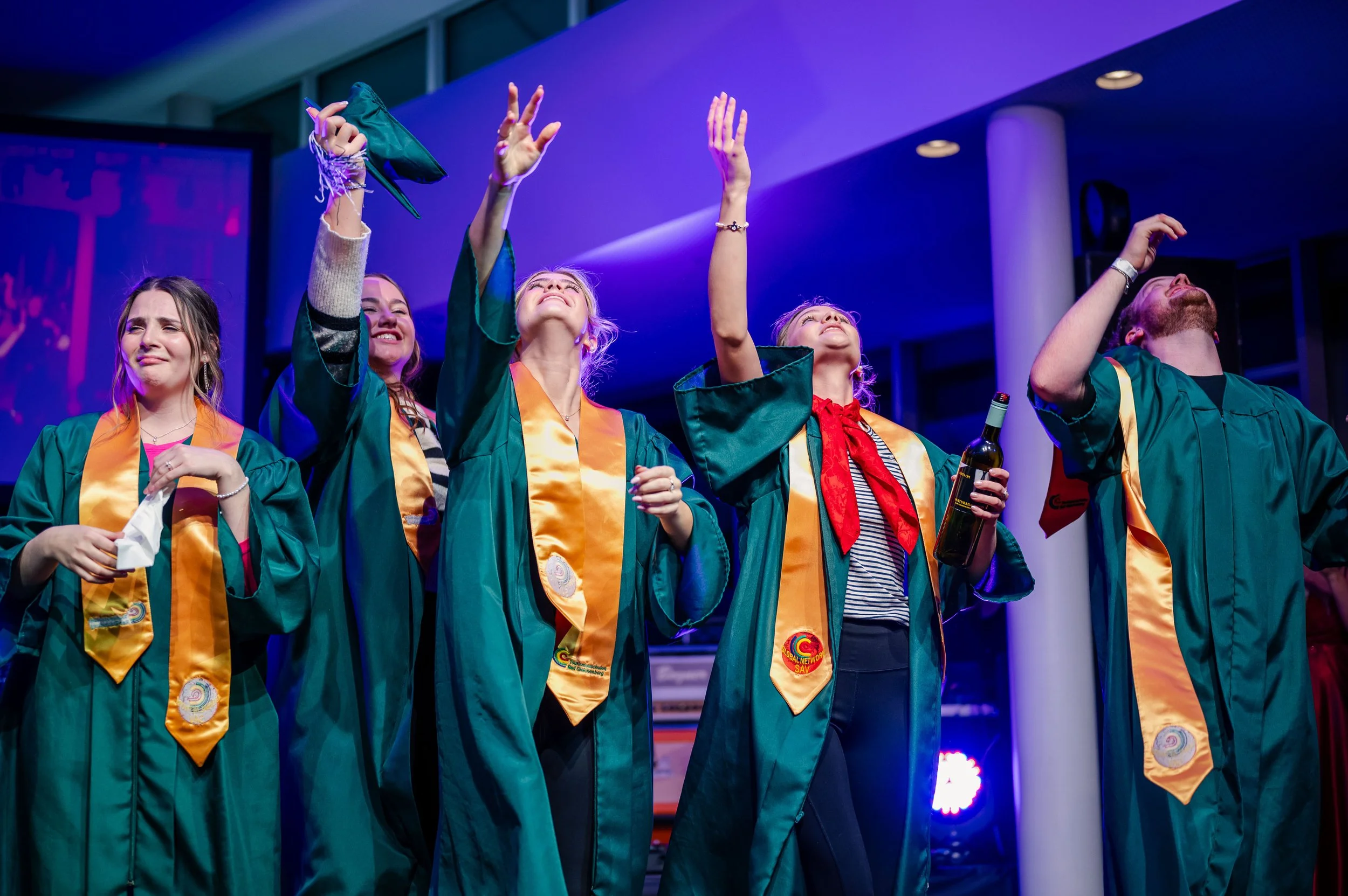 Eine Gruppe von Menschen in Graduate-Roben, die auf einer Bühne feiern, einige lachen und andere blicken nach oben.