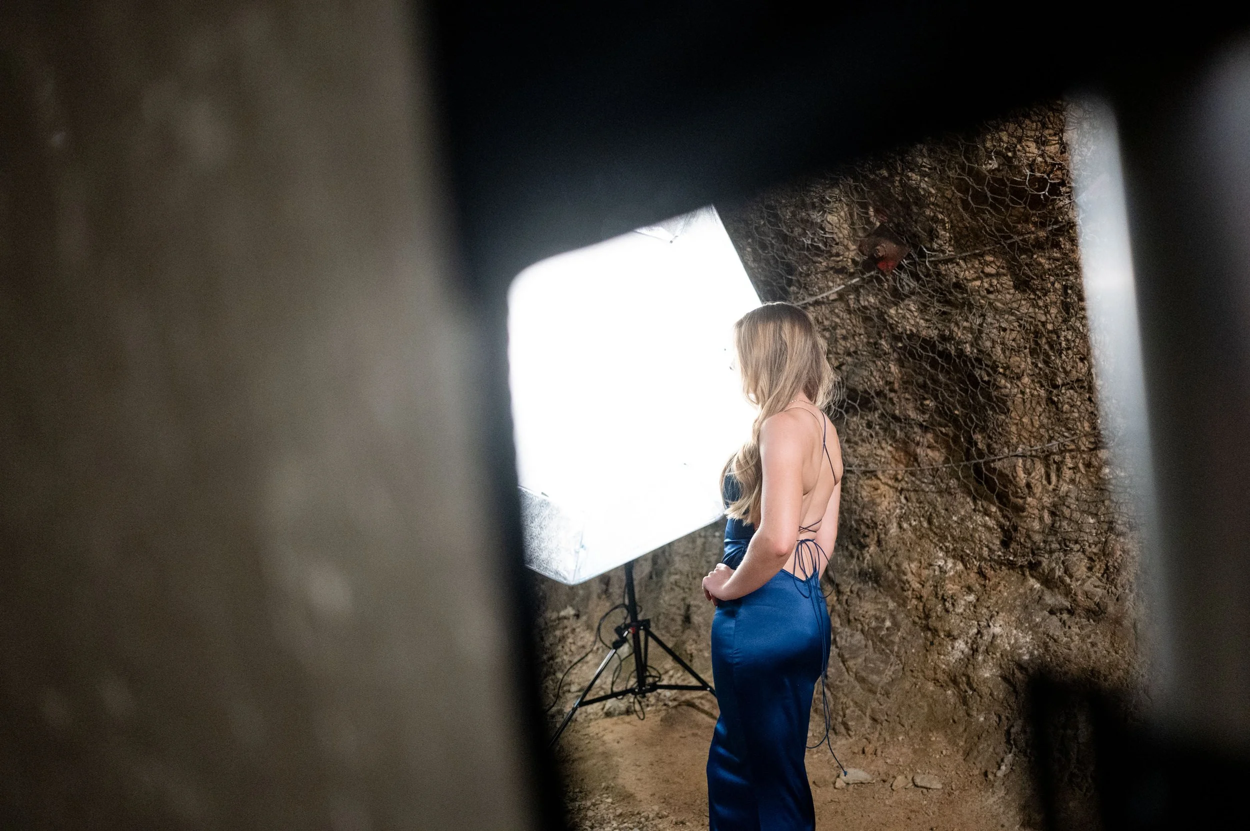 Frau in einem blauen Abendkleid steht vor einem hellen Licht auf einem Gerüst vor einer Backsteinmauer, fotografiert von einer Kamera aus einer dunklen Perspektive.